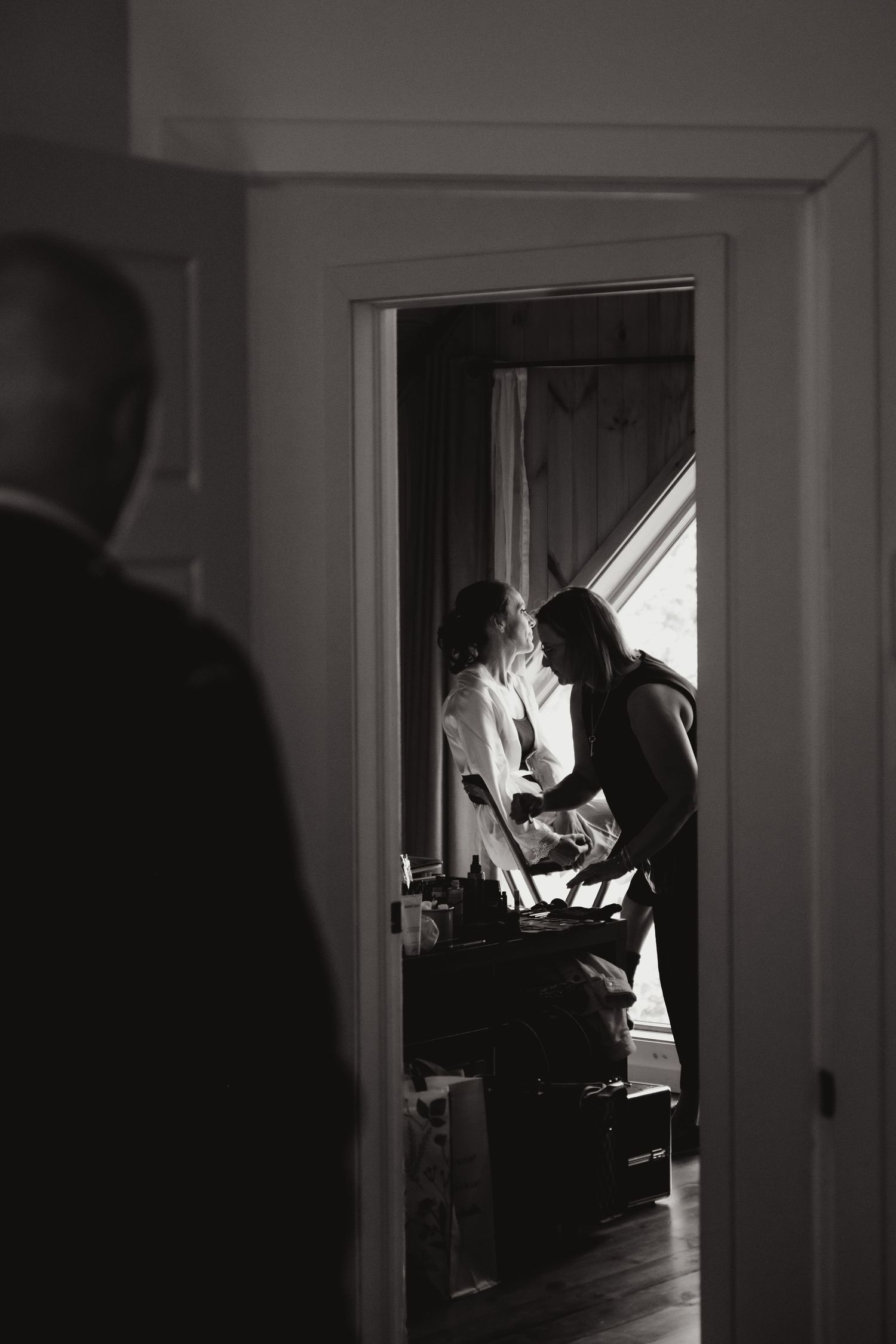 Black and white photo of bride getting ready, viewed through doorway with silhouetted figure in foreground.
