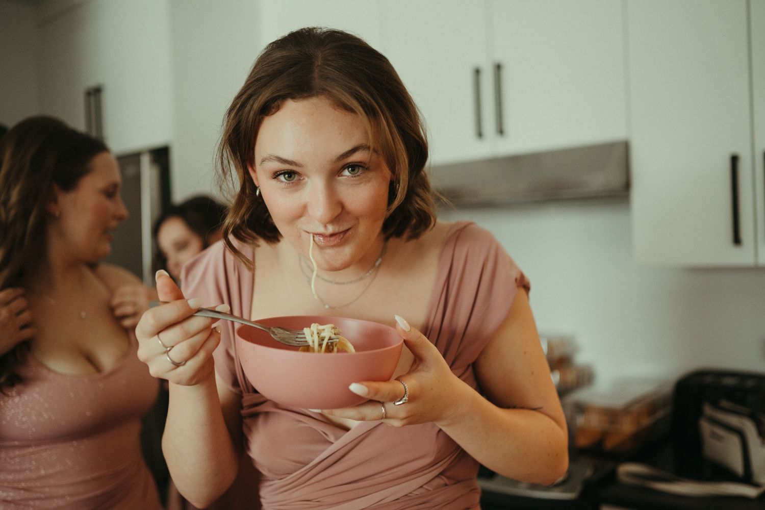 Bridesmaid in blush pink dress eating from a bowl in a kitchen before a wedding.