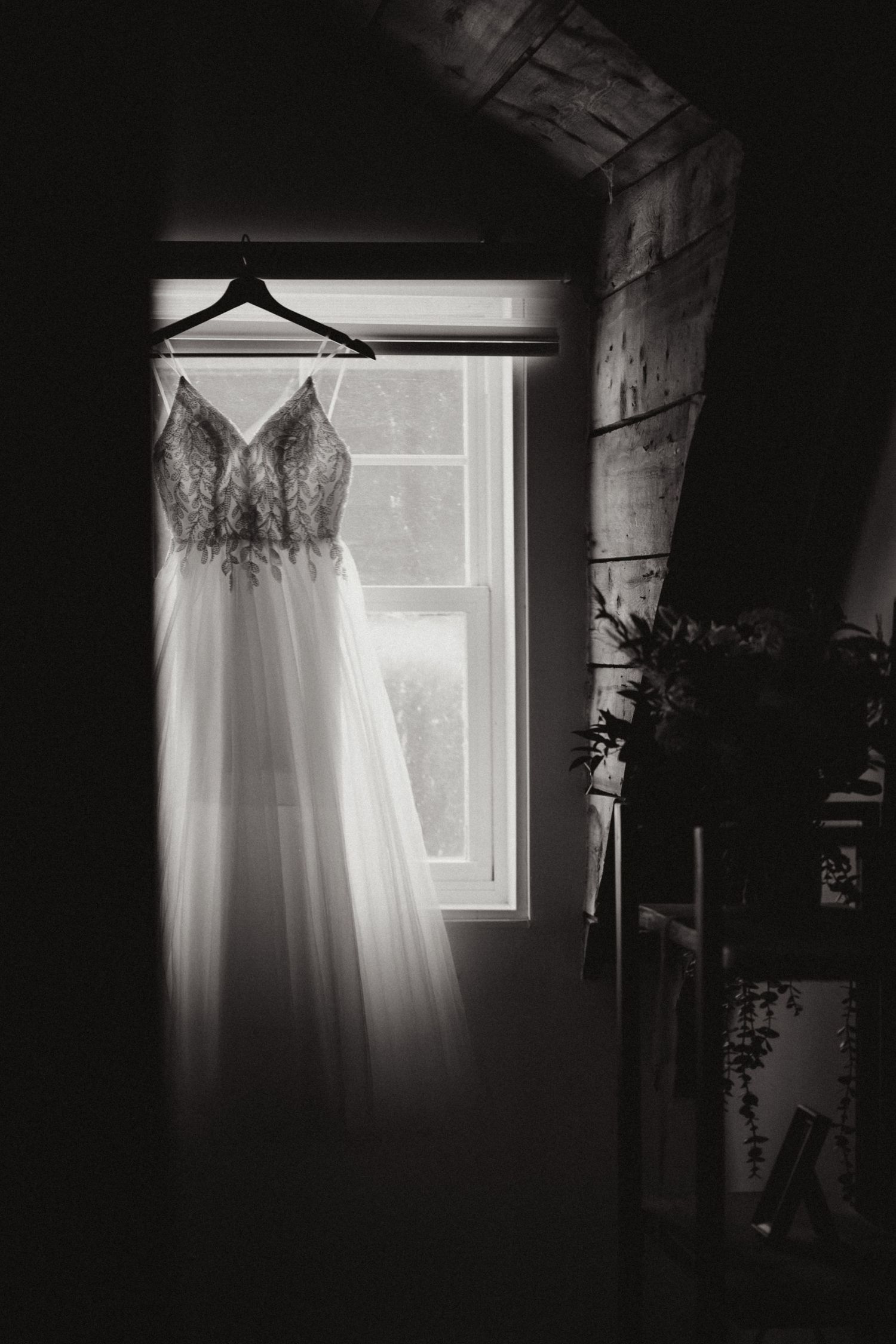 An elegant lace wedding dress hangs on a hanger before a window in a black and white photo.