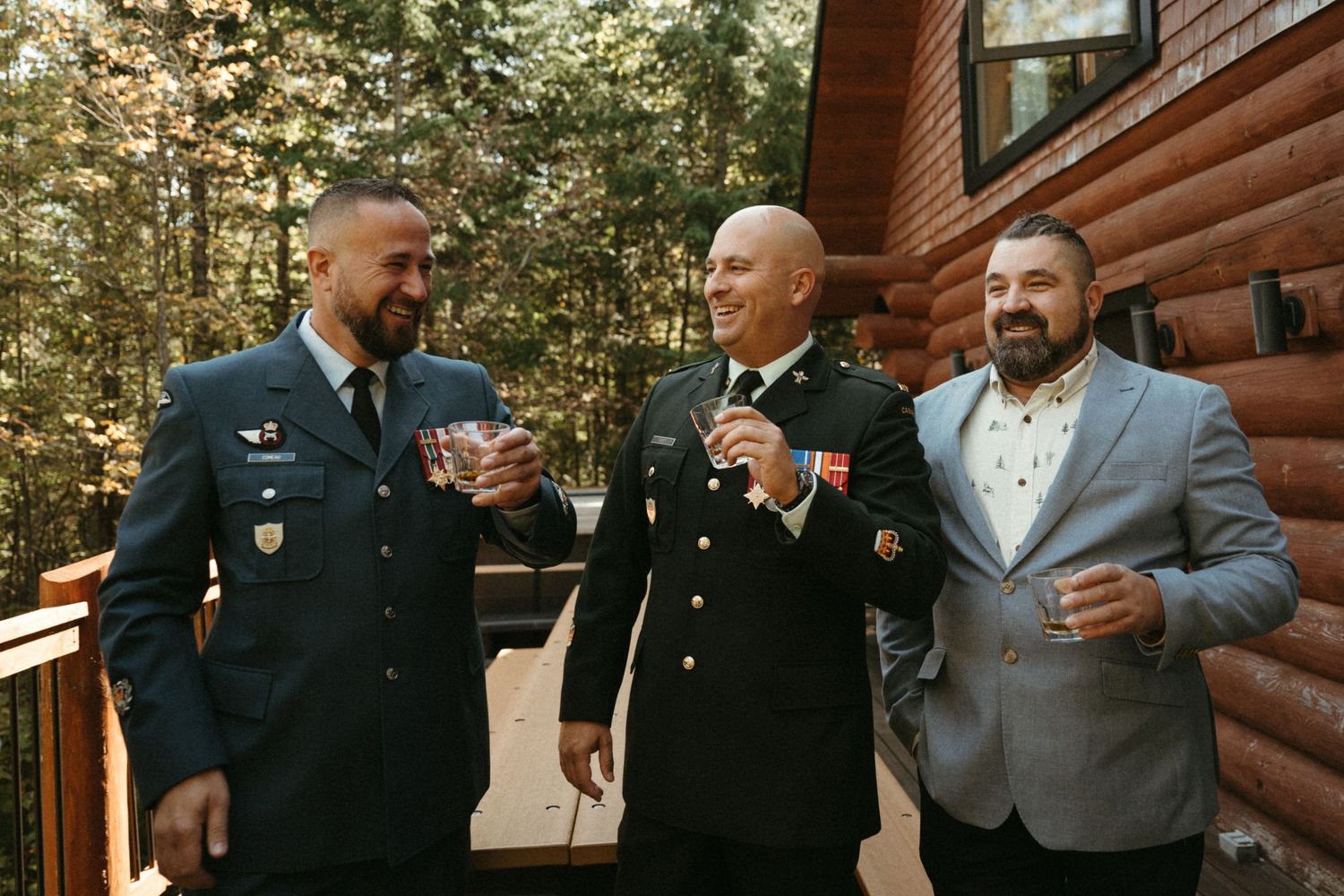 Three men in military uniforms and suits toasting with drinks on a wooden cabin deck surrounded by trees.