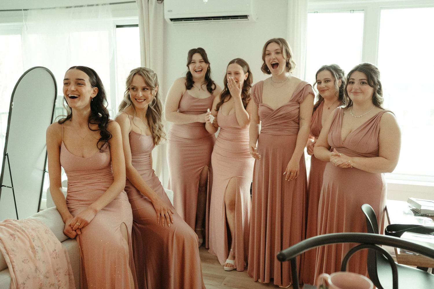 Six bridesmaids in dusty rose gowns reacting with joy and surprise at a wedding.
