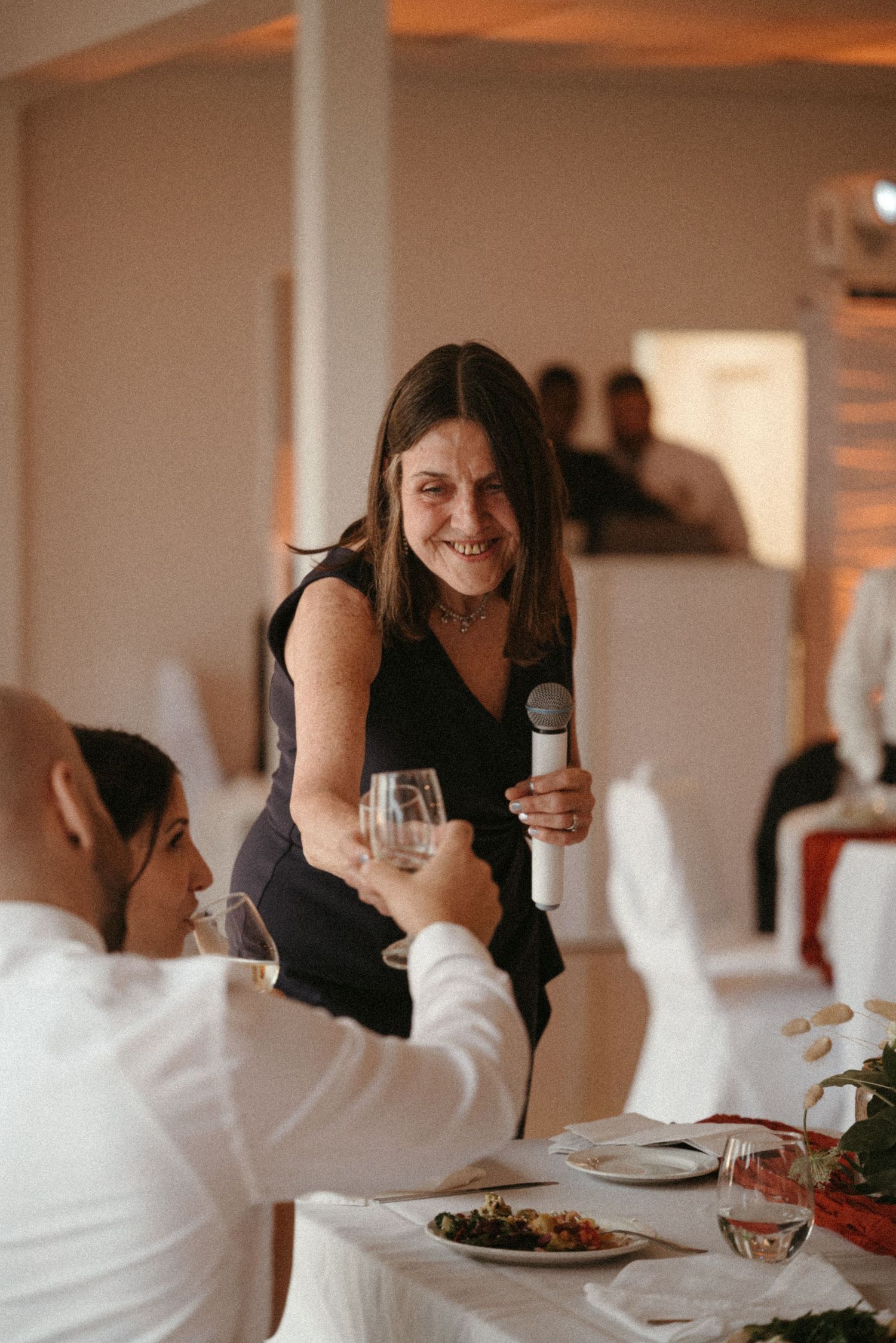 Woman in black dress giving a toast speech with microphone and wine glass at wedding reception.