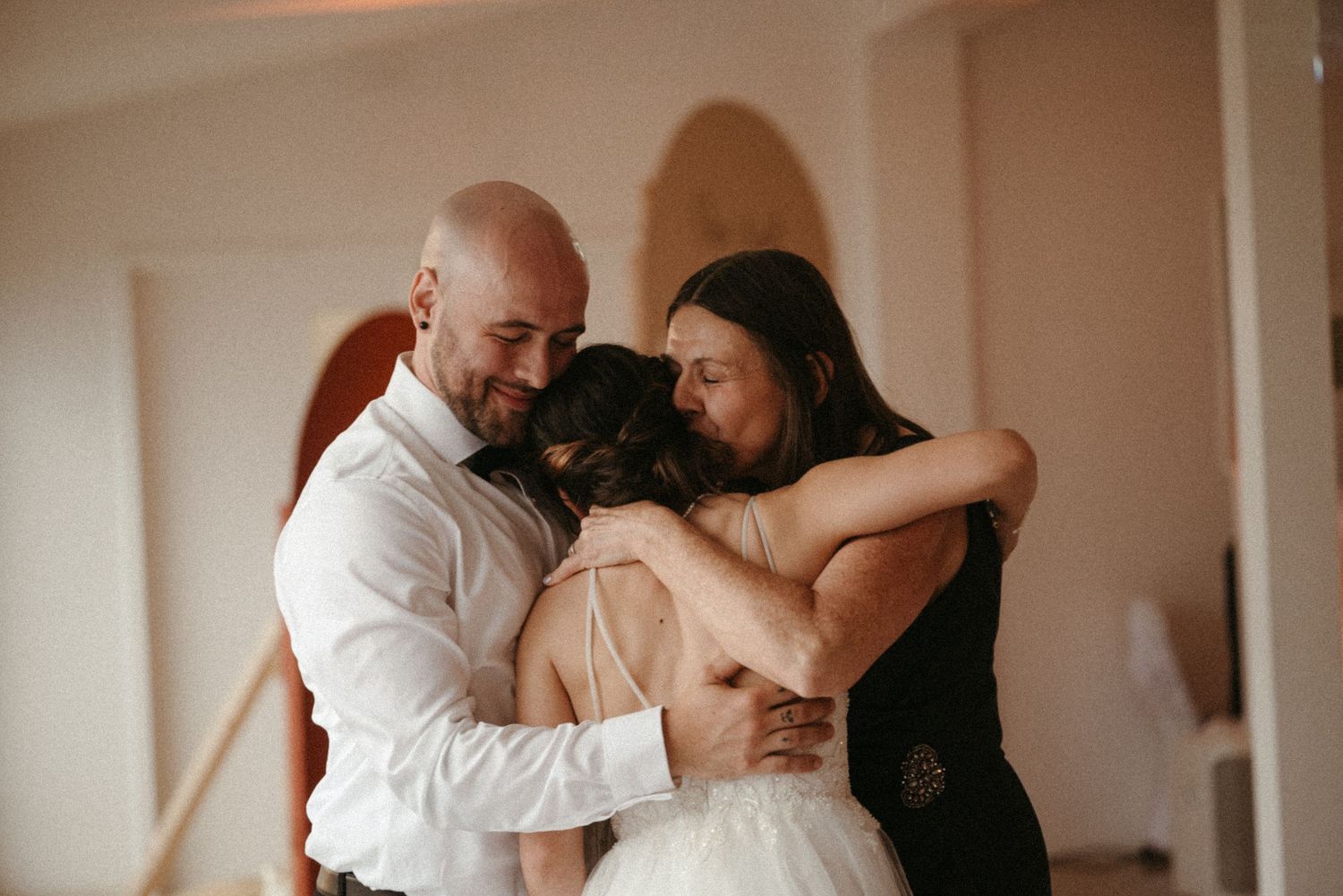 Bride and groom share emotional group hug with wedding guest in elegant venue during special celebration.