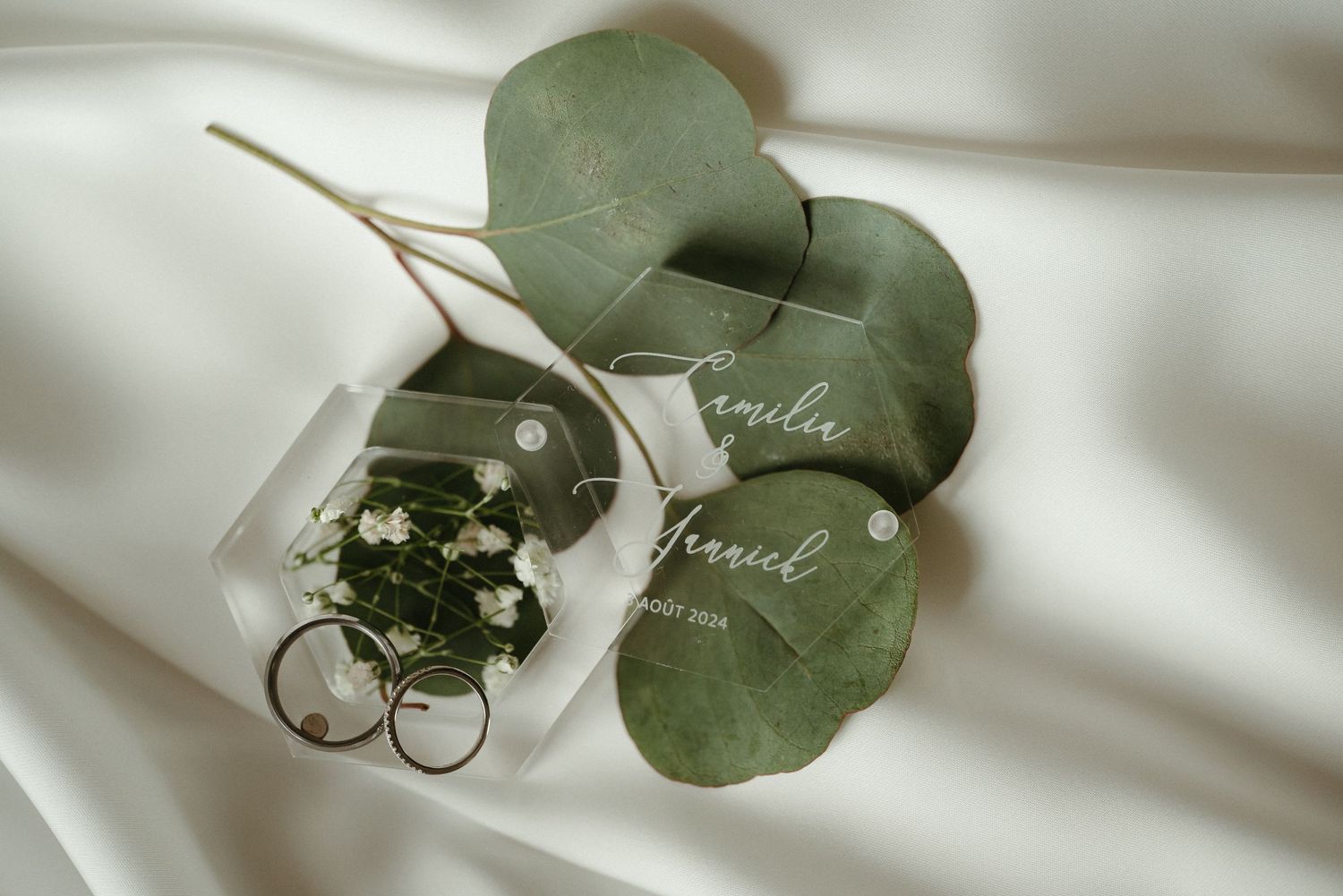 Wedding rings and acrylic name card with eucalyptus leaves on white satin fabric.