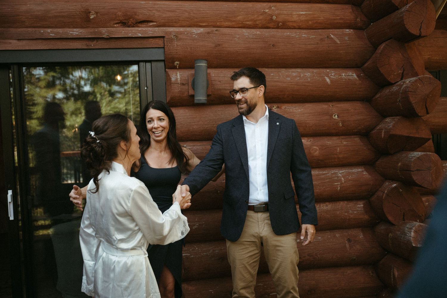 Man and woman shaking hands near a log cabin entrance while another woman smiles in the background.