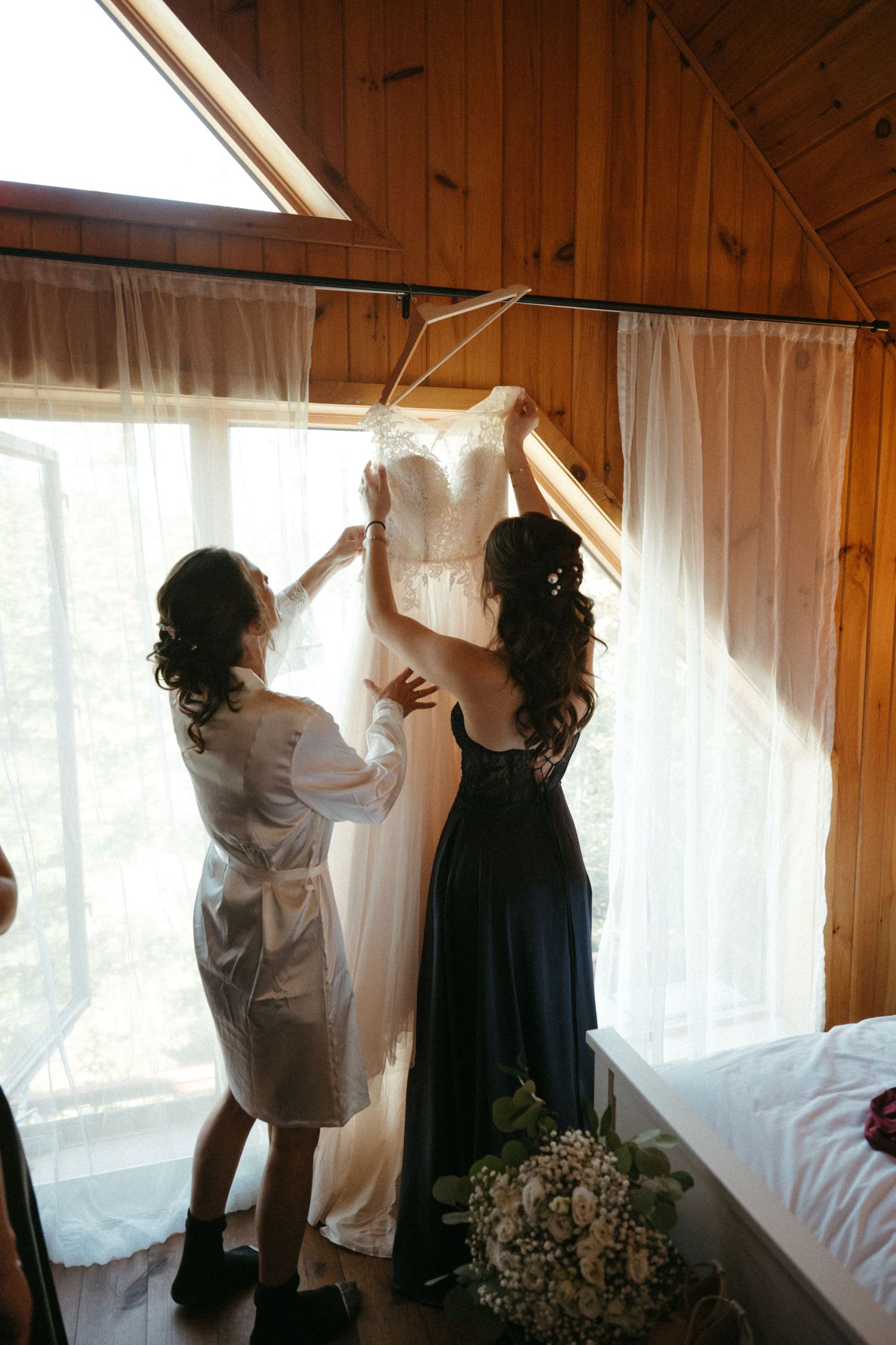 Two women lifting a white wedding dress near a sunlit window in a wooden cabin bedroom.
