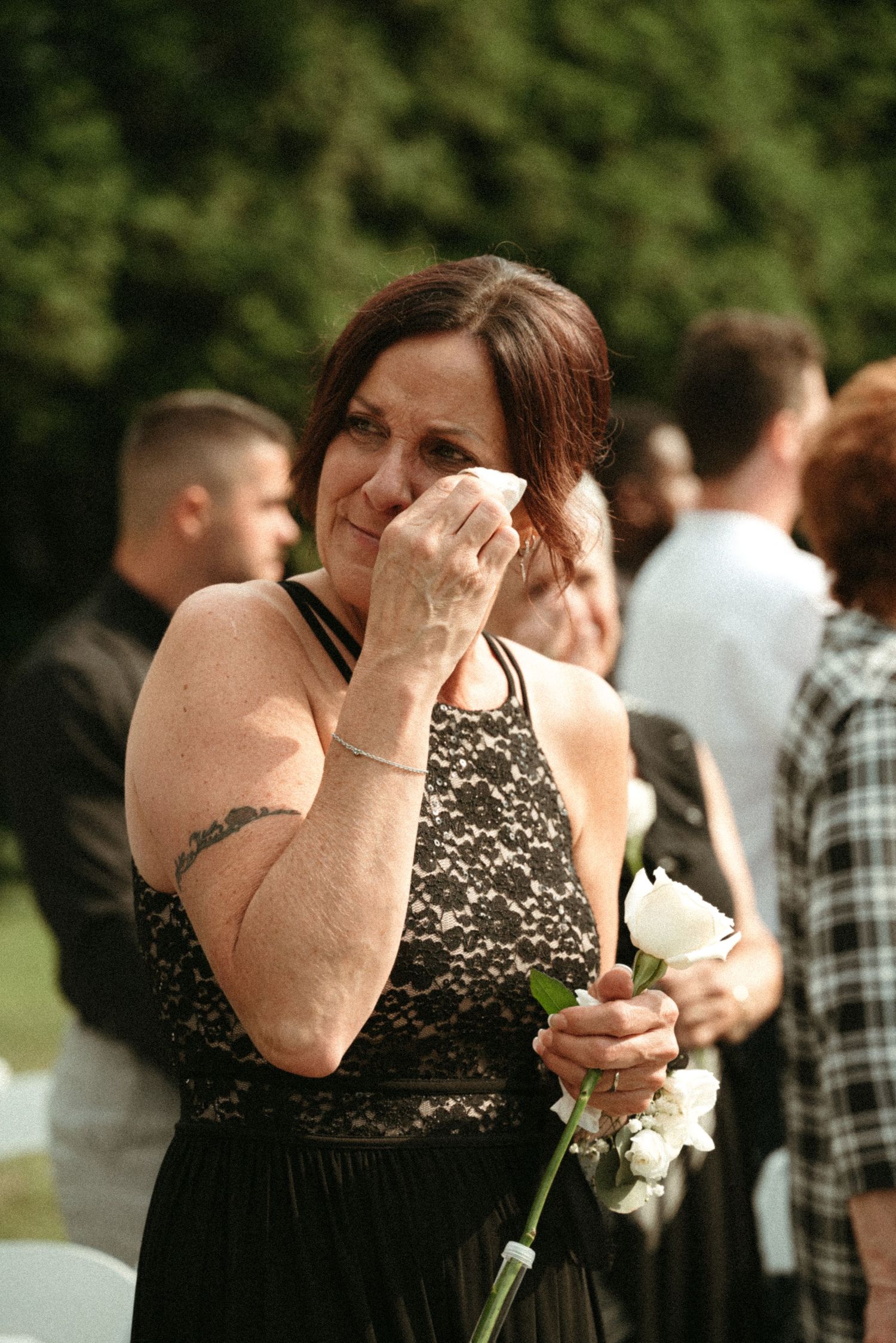 Emotional wedding guest wiping tears while holding white rose during outdoor ceremony.