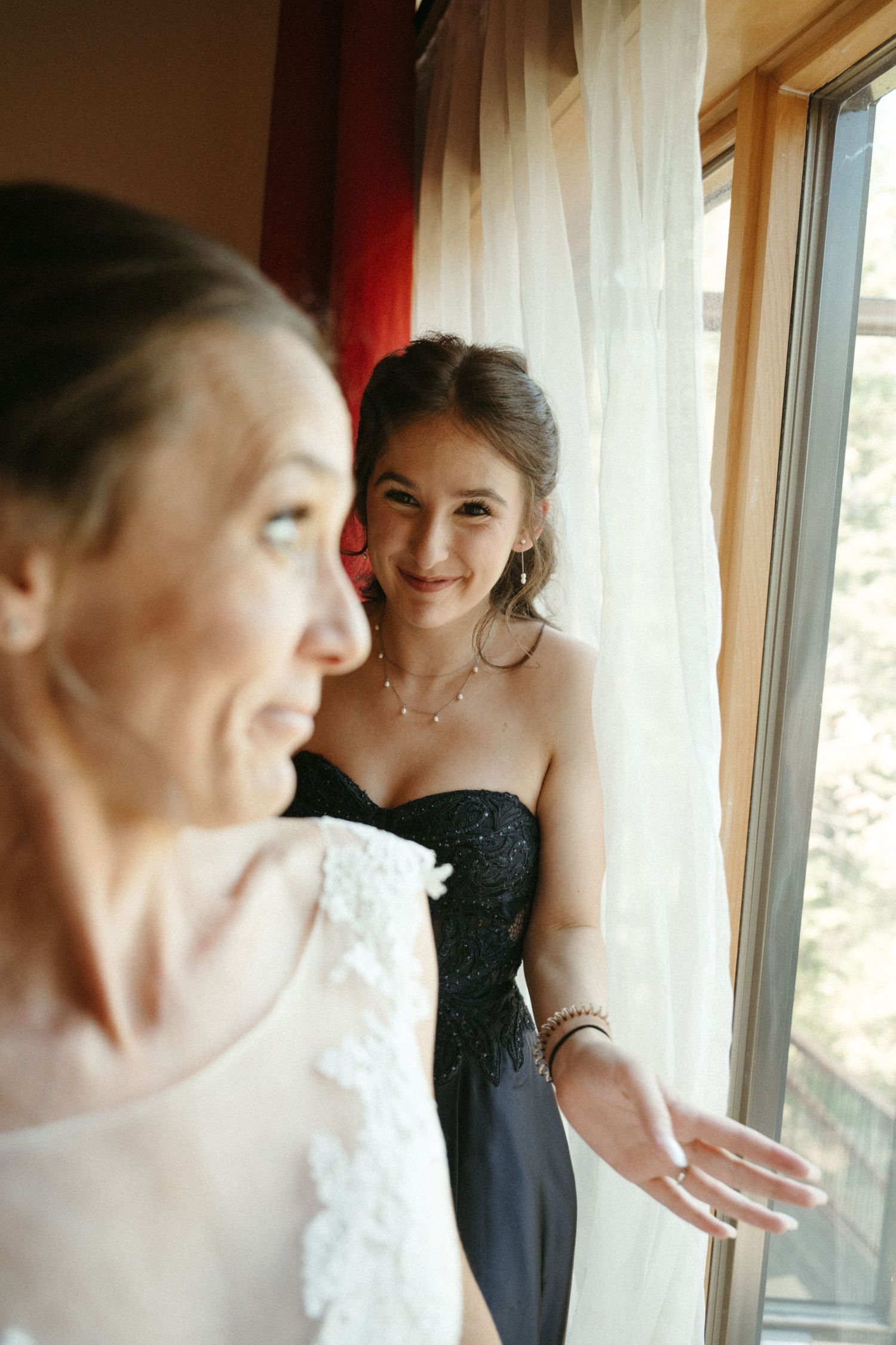 Bridesmaid in black strapless gown smiles at bride in white lace dress near a curtained window.