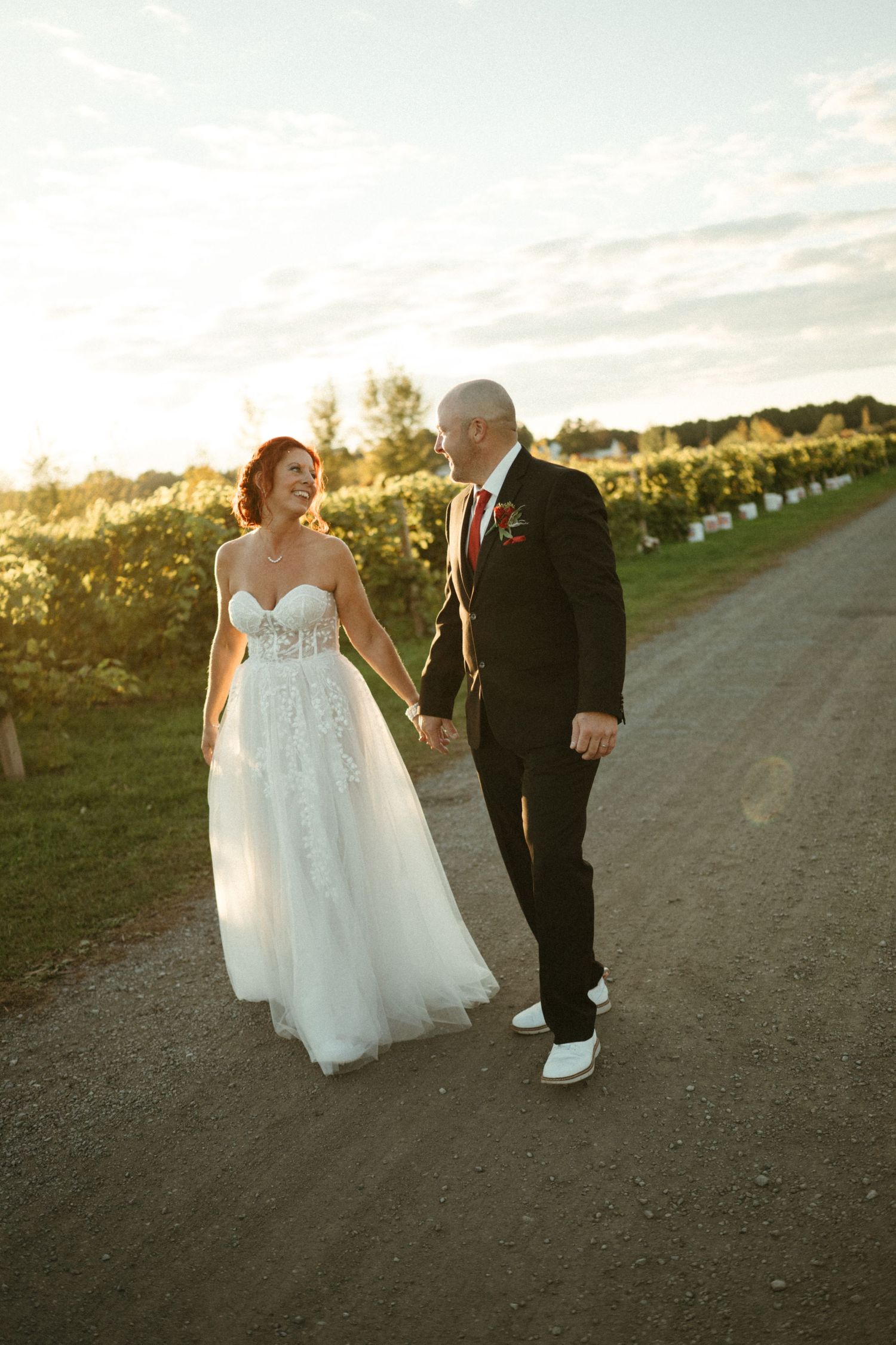 Bride and groom walk hand-in-hand along a sunlit vineyard path, smiling at each other in wedding attire.