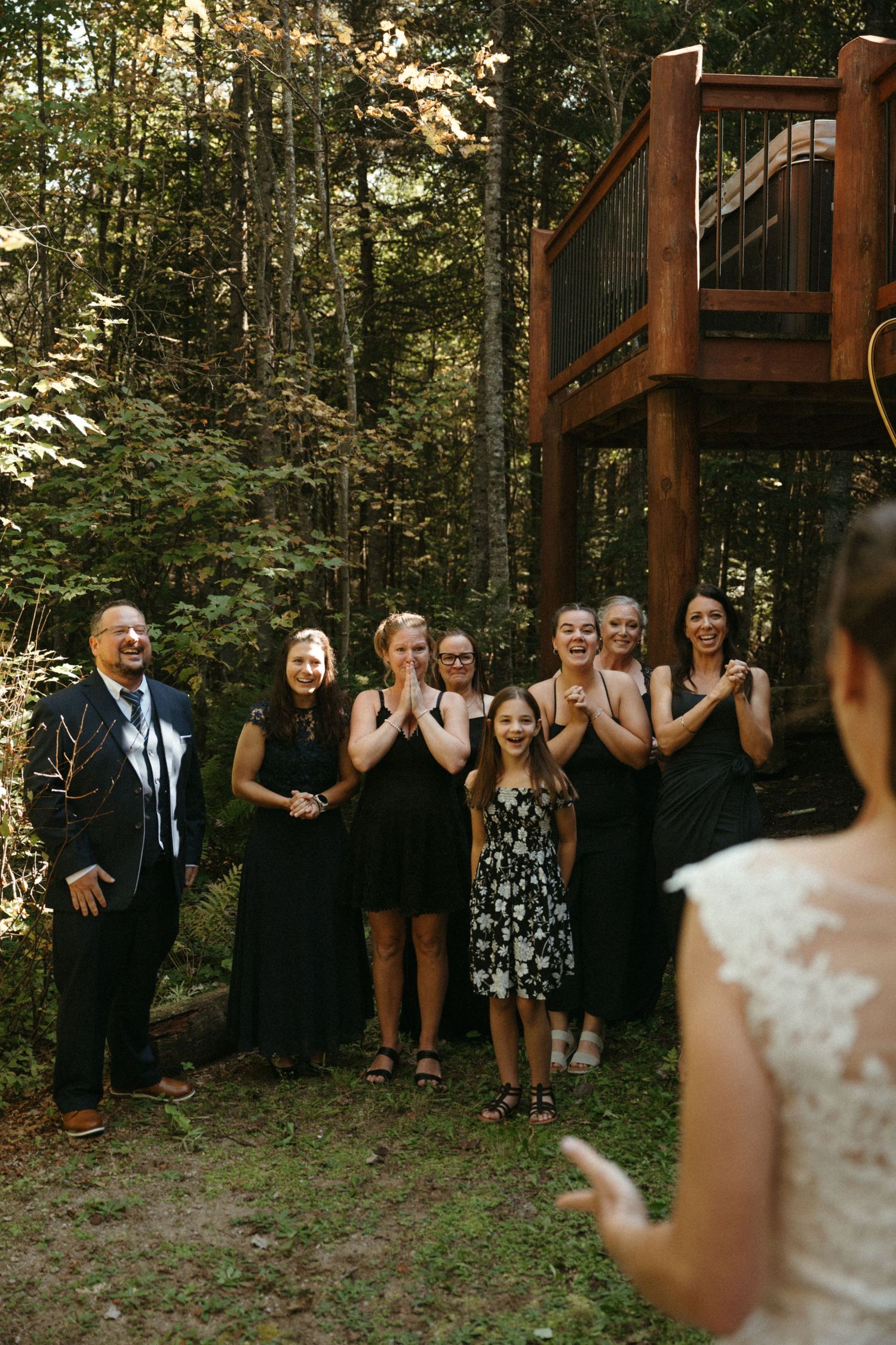 Wedding guests in black attire reacting with joy upon seeing the bride during a forest first look reveal.