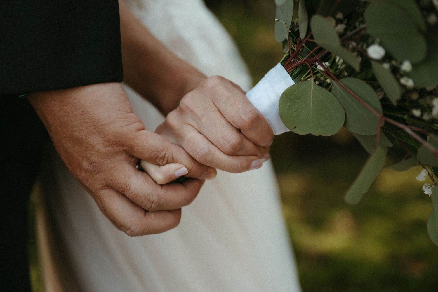 Close-up of bride and groom holding hands during wedding ceremony with bridal bouquet of eucalyptus.