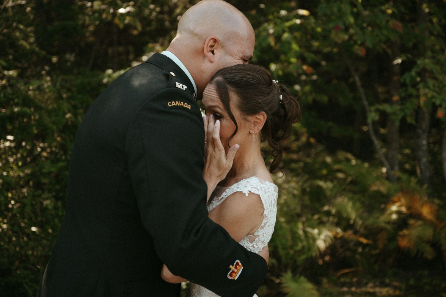 Emotional military groom in Canadian Army uniform gently wiping tears from his bride's face in a forest setting.