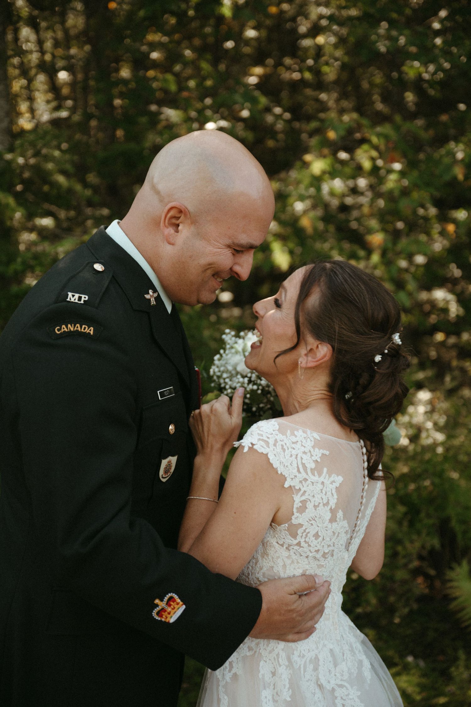 Military groom in Canadian uniform shares tender moment with bride in lace wedding dress outdoors.