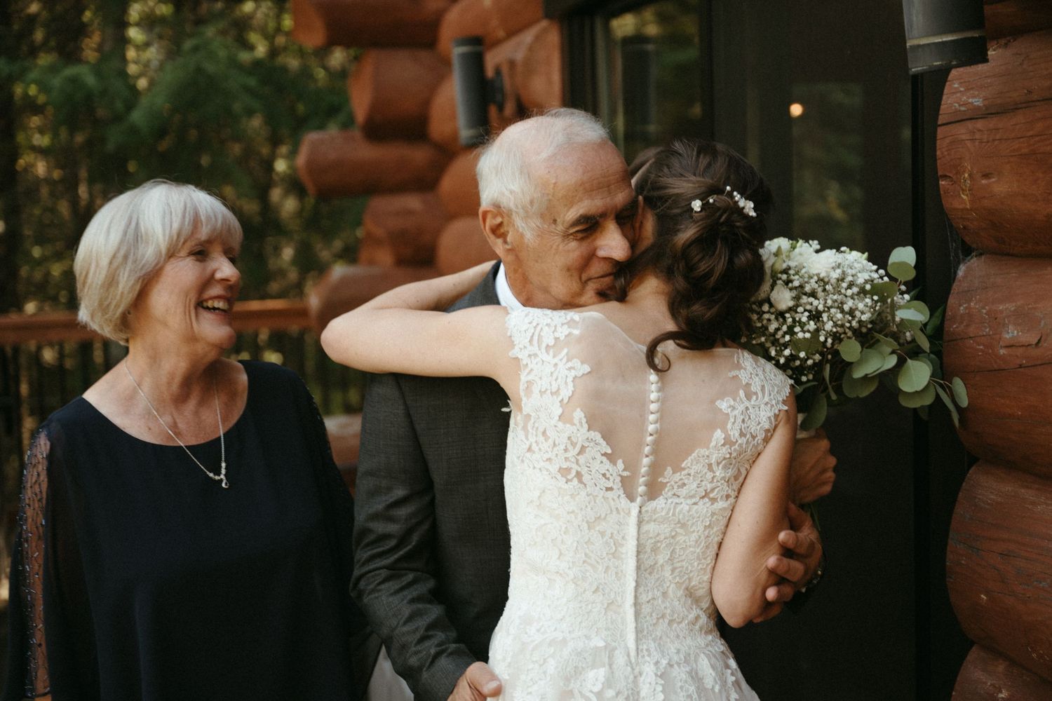 Bride in lace gown embraces older man while woman smiles beside log cabin venue.