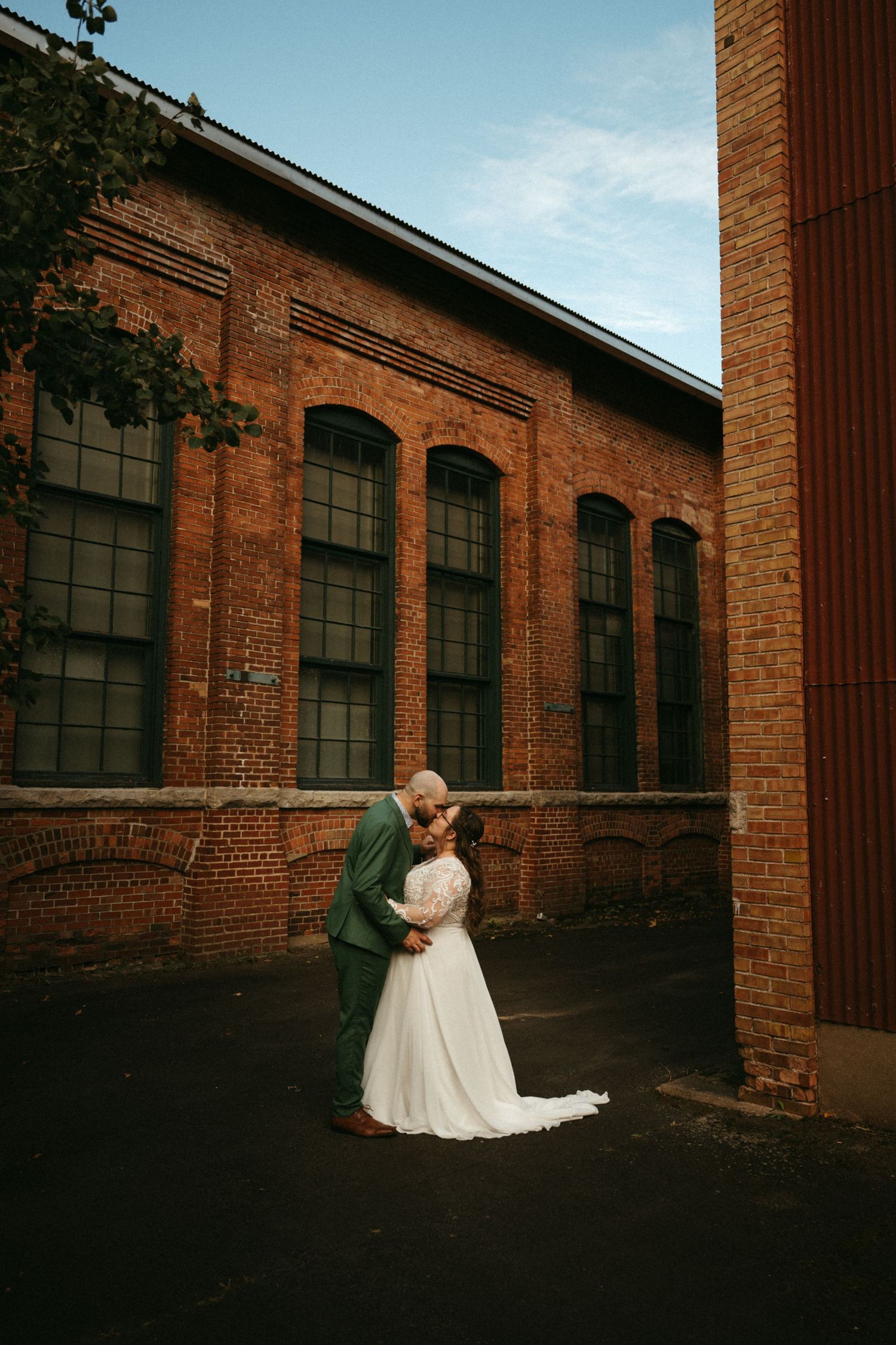 Bride and groom share a romantic kiss outside a rustic brick building on their wedding day.
