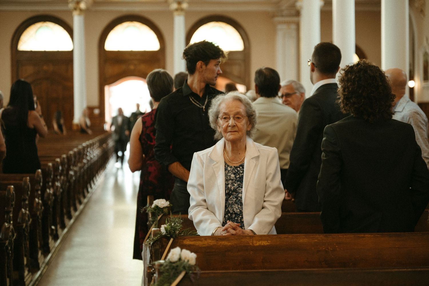 Elderly woman in white blazer sits in church pew smiling during wedding ceremony with guests gathered behind her.
