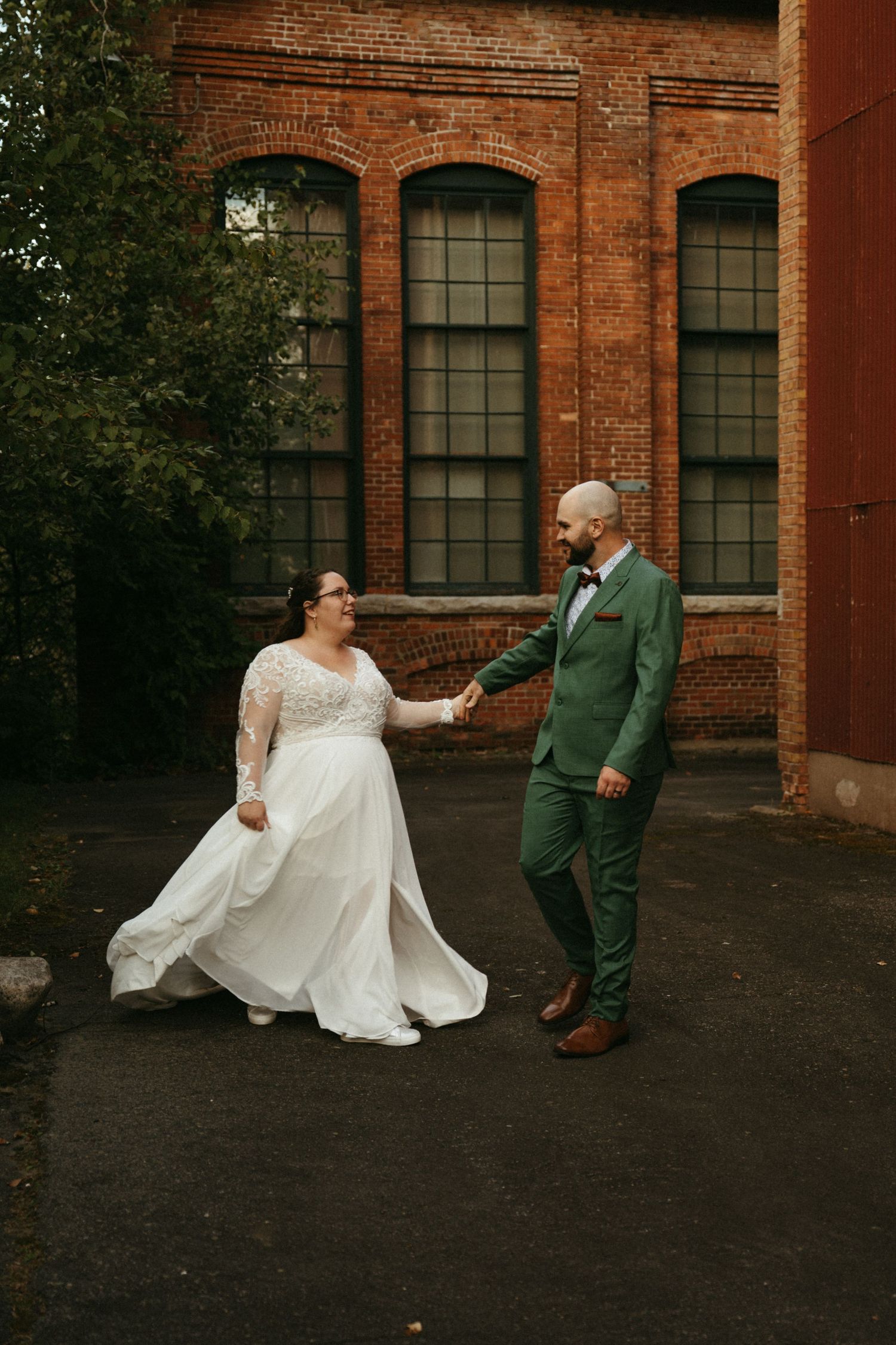 Bride in white lace gown and groom in green suit hold hands outside a rustic brick building on their wedding day.
