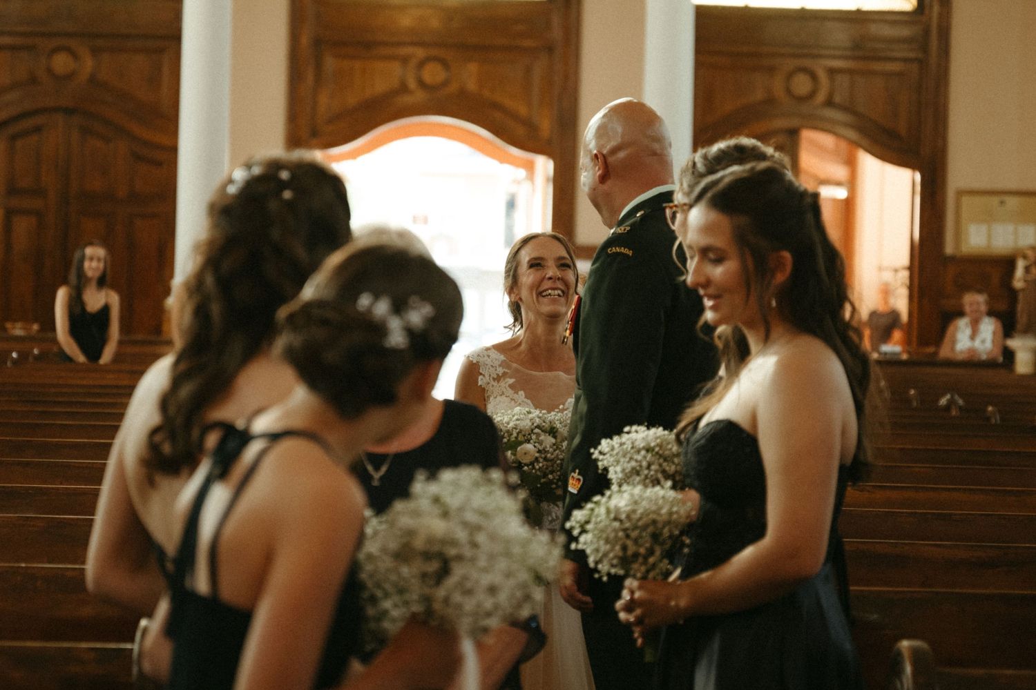 Bride laughs with bridesmaids in black dresses holding baby's breath bouquets inside historic church.