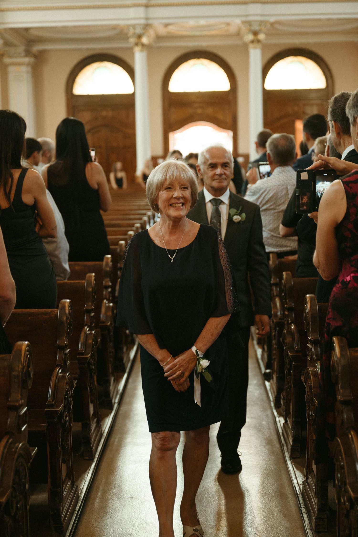 Elegant woman in black dress walks down church aisle with man in suit during wedding ceremony.