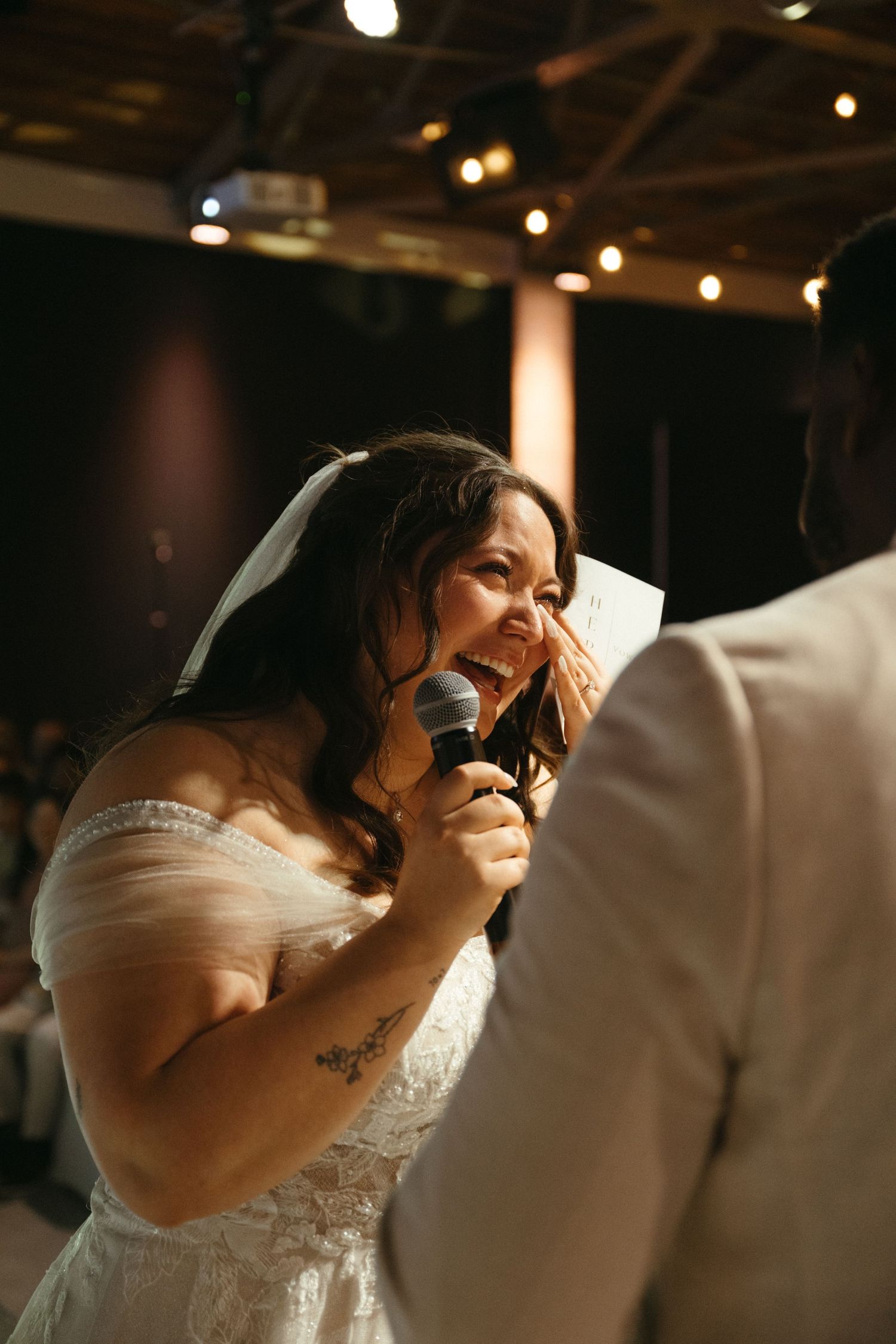 Emotional bride in white lace dress speaking into microphone during wedding reception vows.