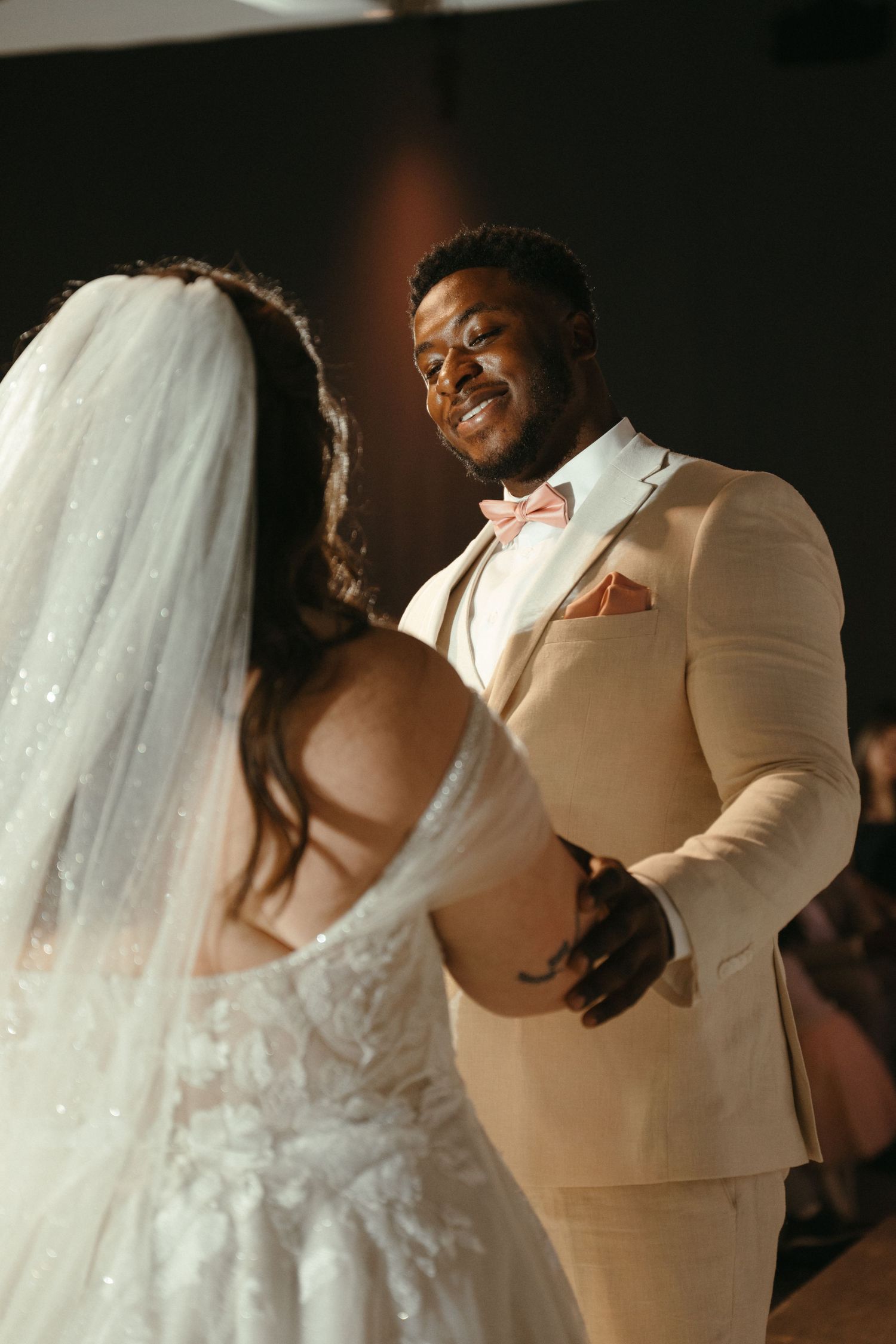 Groom in beige suit and pink bow tie smiles at bride during their first dance at wedding reception.