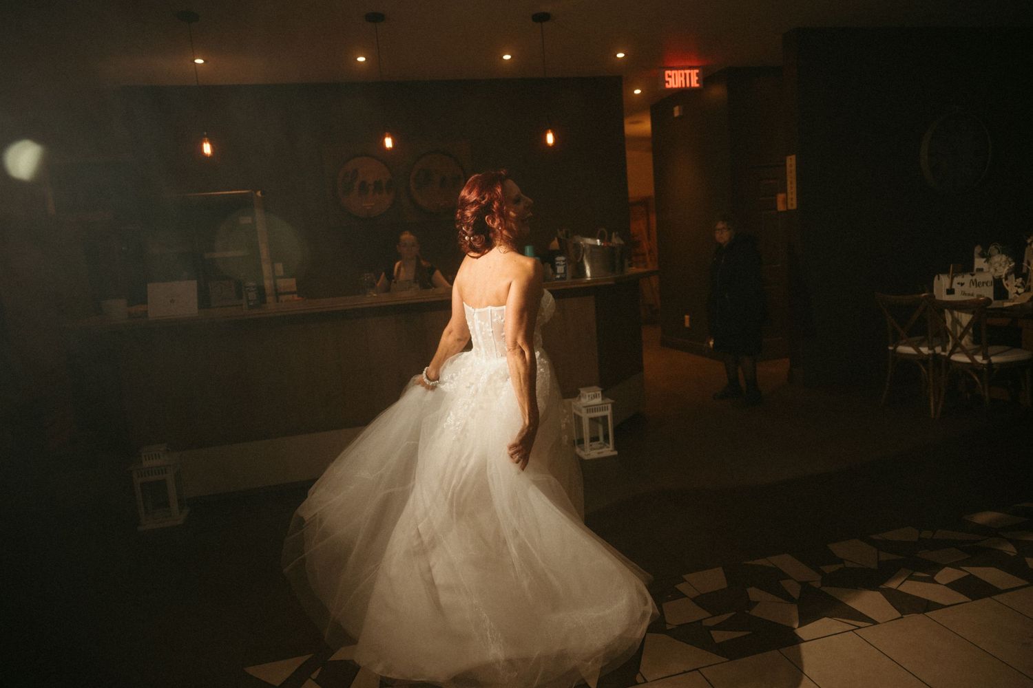 Bride in a flowing white ball gown twirling in a dimly lit wedding venue with checkered floors.