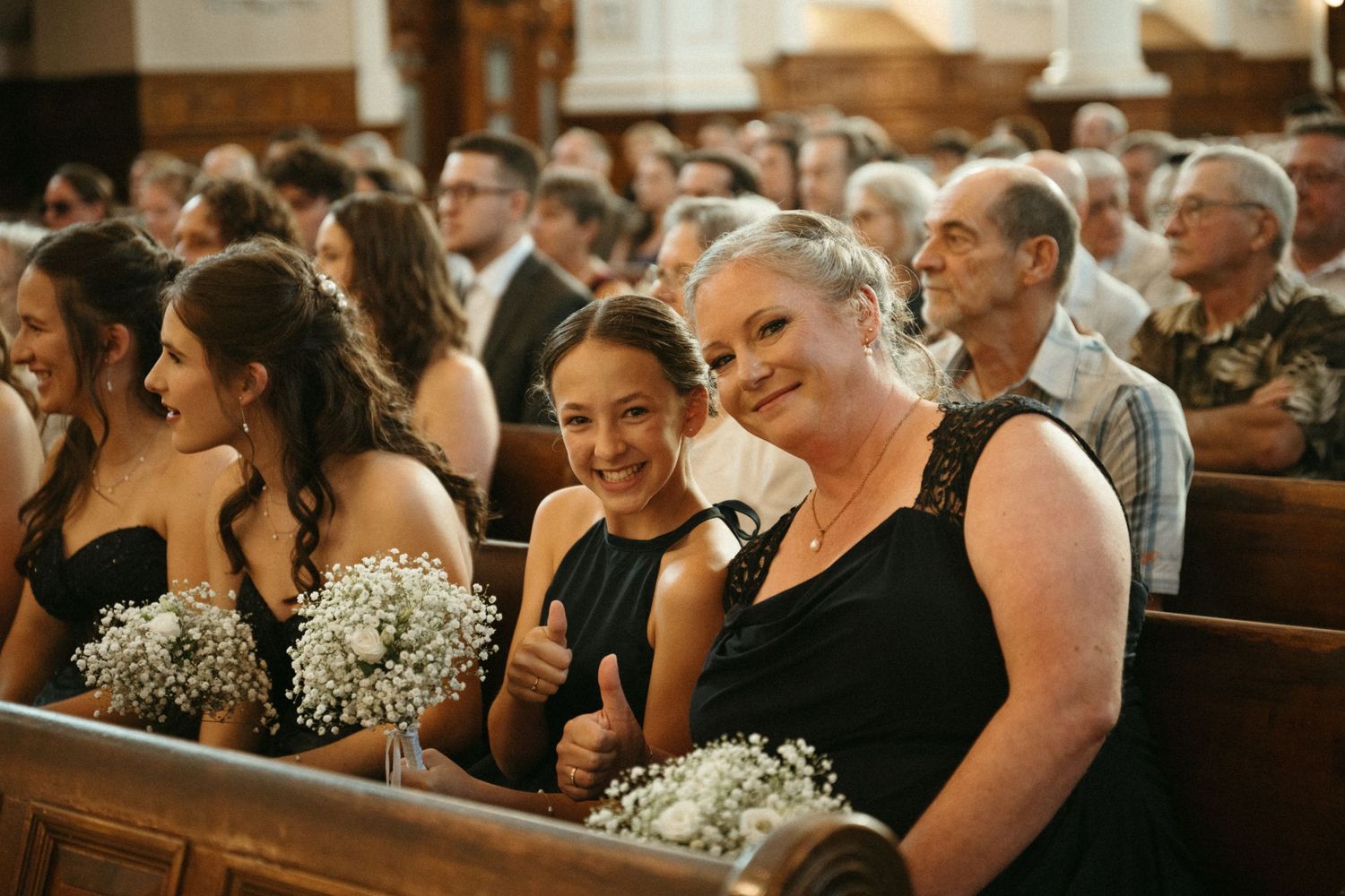 Women in black dresses holding white floral bouquets smiling in a church pew during a wedding ceremony.