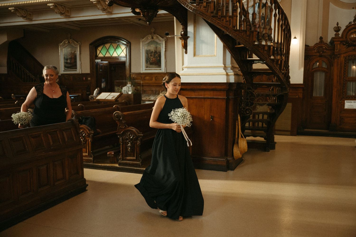 Bridesmaid in black gown walking through ornate wooden church interior before wedding ceremony.