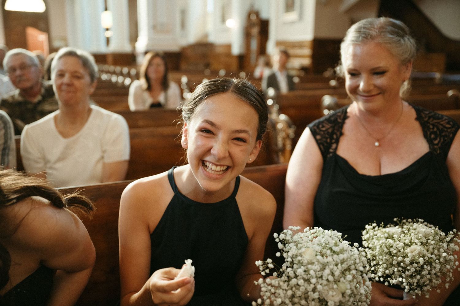 Joyful bridesmaid in black dress laughing while holding baby's breath bouquet during wedding ceremony.