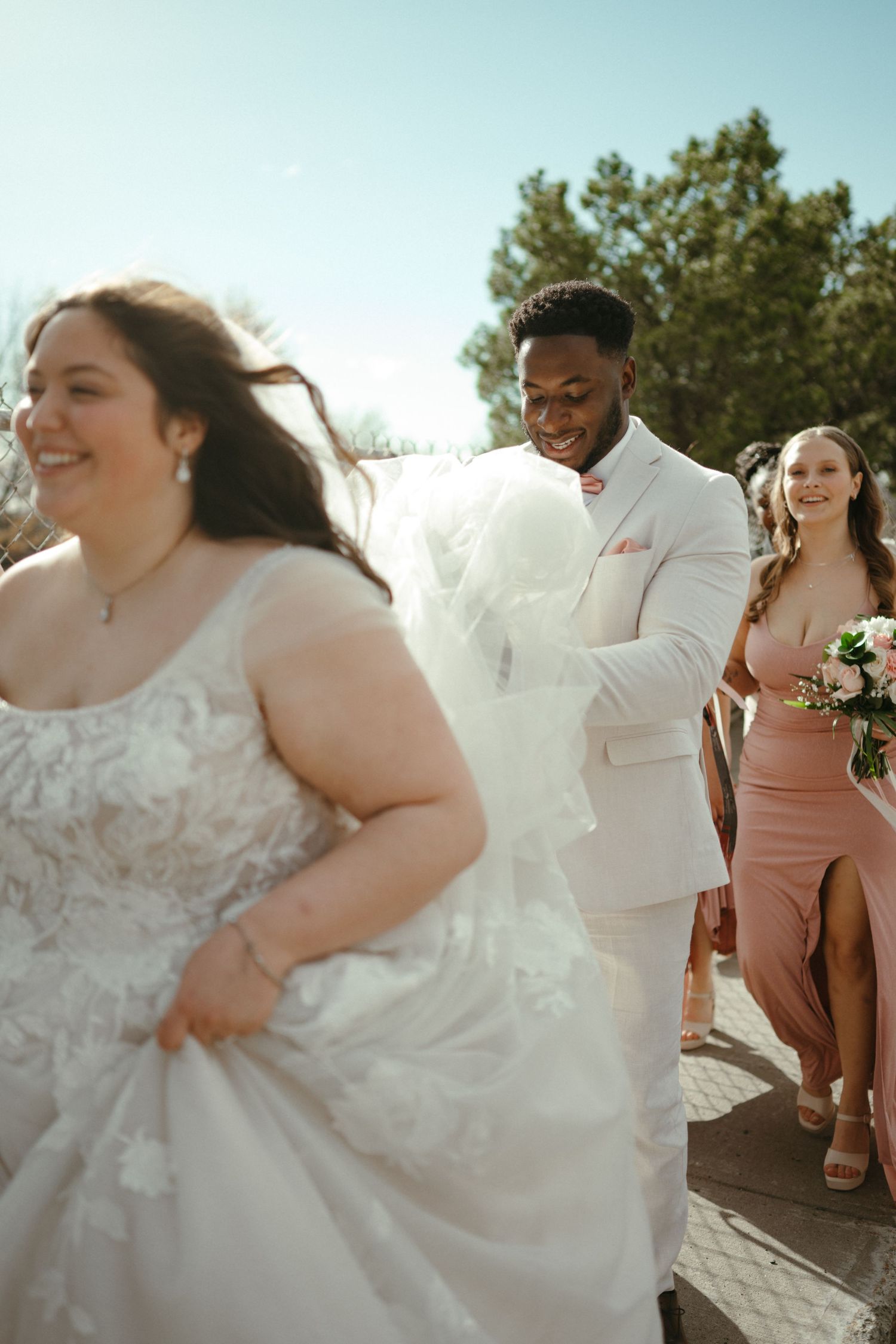 Smiling groom in white suit walking with bride in lace gown and bridesmaids outdoors.