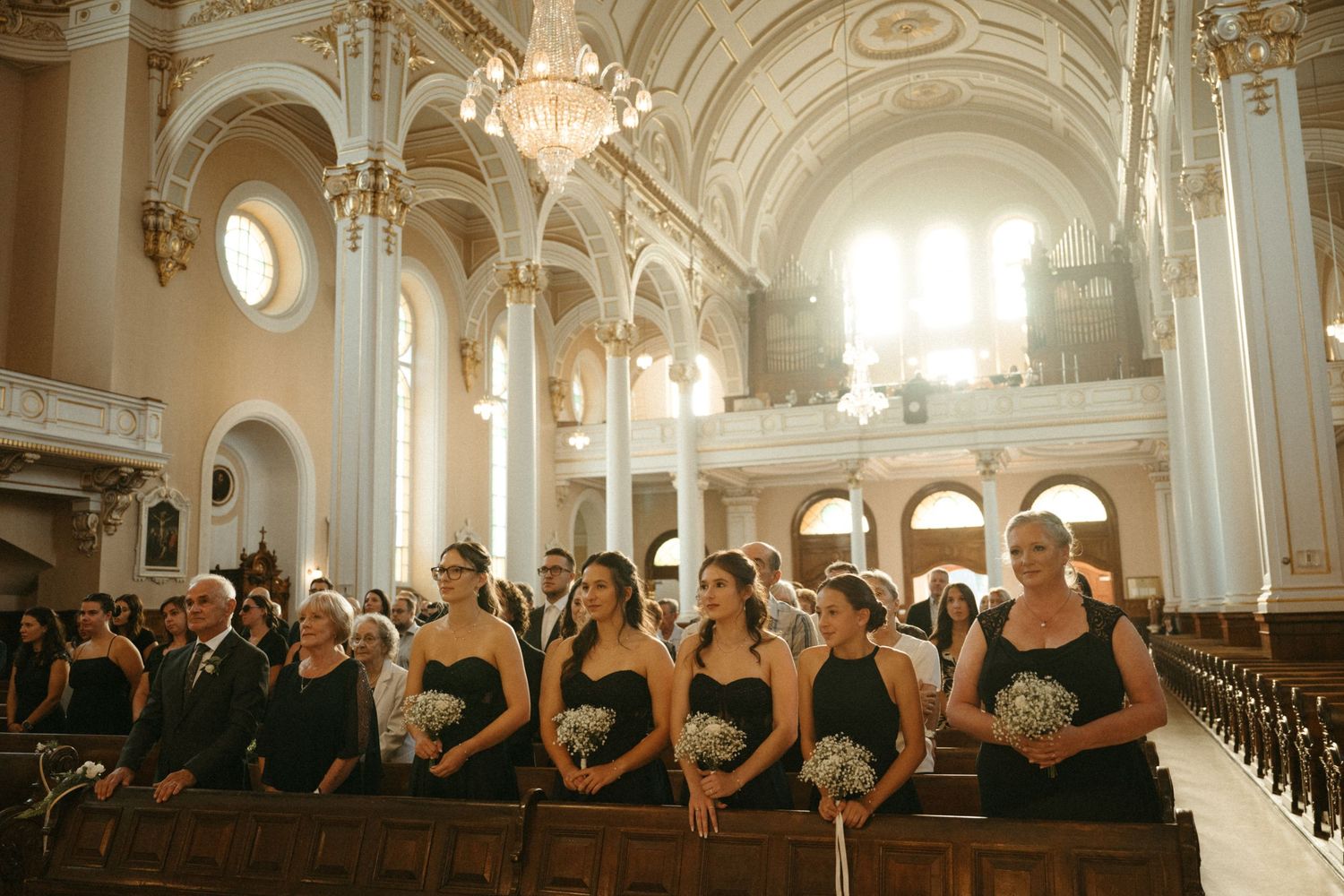 Bridesmaids in black strapless gowns holding white bouquets seated in grand cathedral church pews.