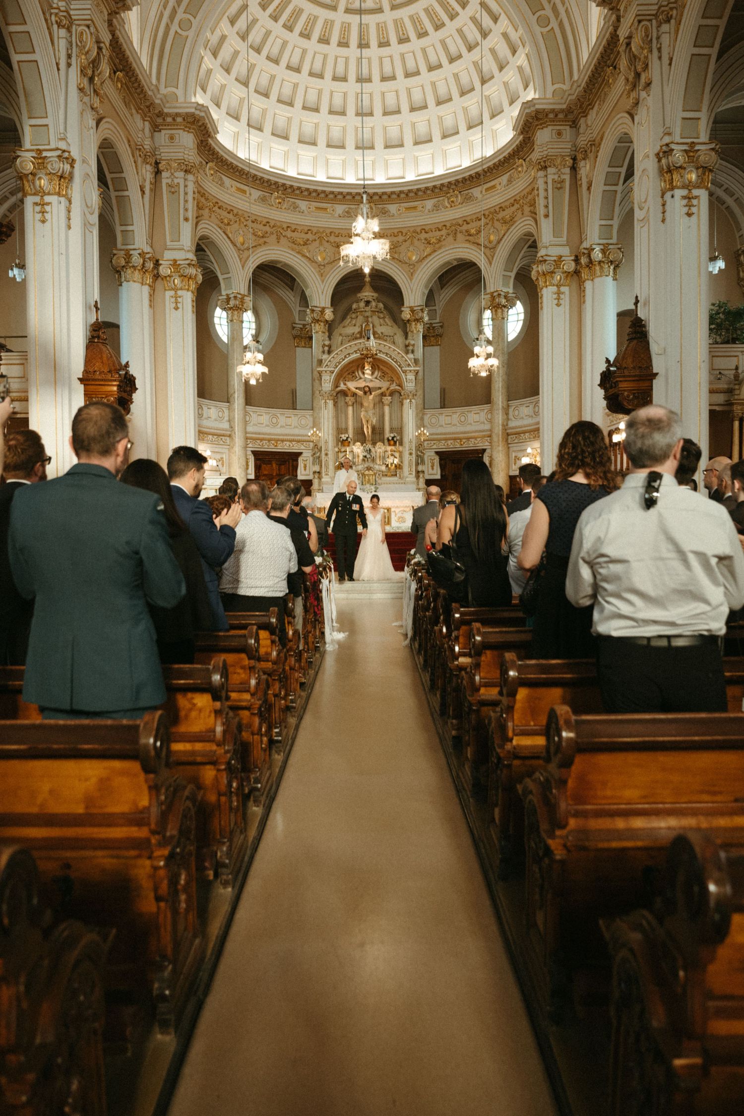 Grand cathedral wedding ceremony with bride and groom at ornate altar surrounded by seated guests.