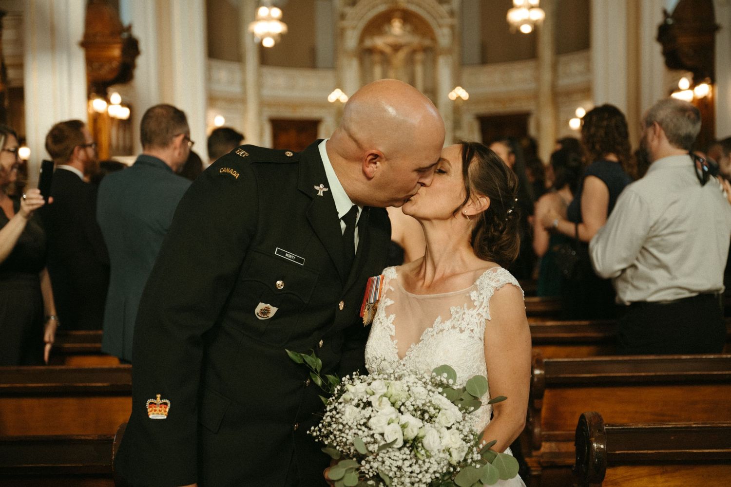 Newlyweds kissing in church aisle as groom holds bride in military uniform with bridal bouquet of white flowers.