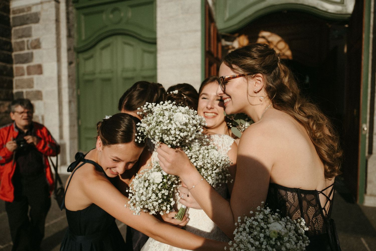 Bride in white gown joyfully embraces bridesmaids in black dresses outside church doors.