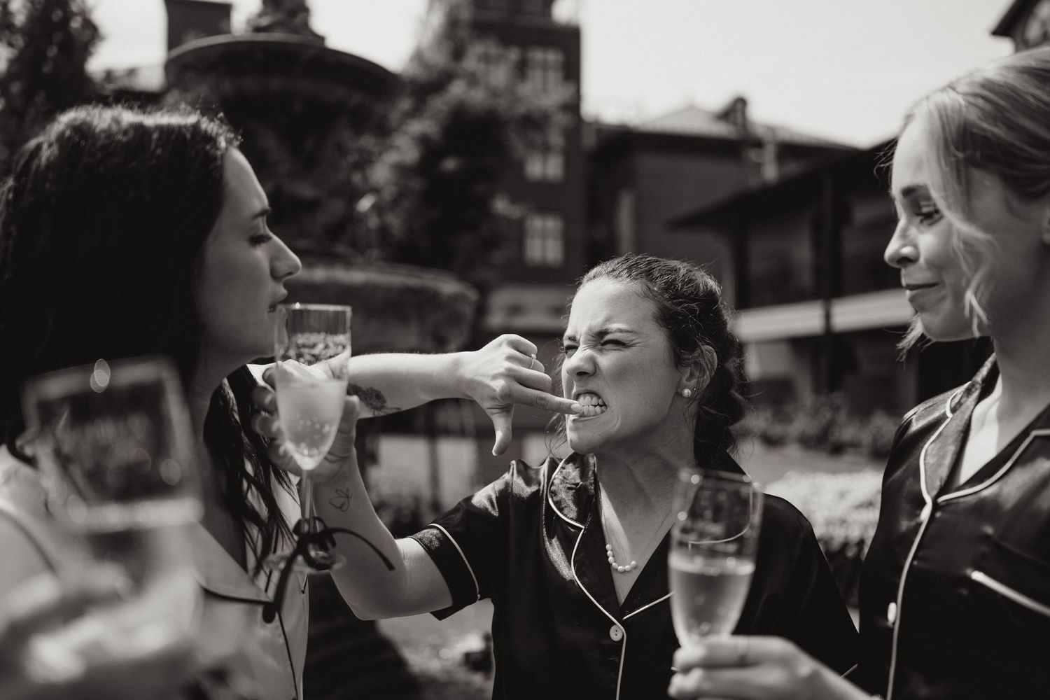 Bridesmaids in satin pajamas laughing and sharing a moment with champagne glasses outdoors.