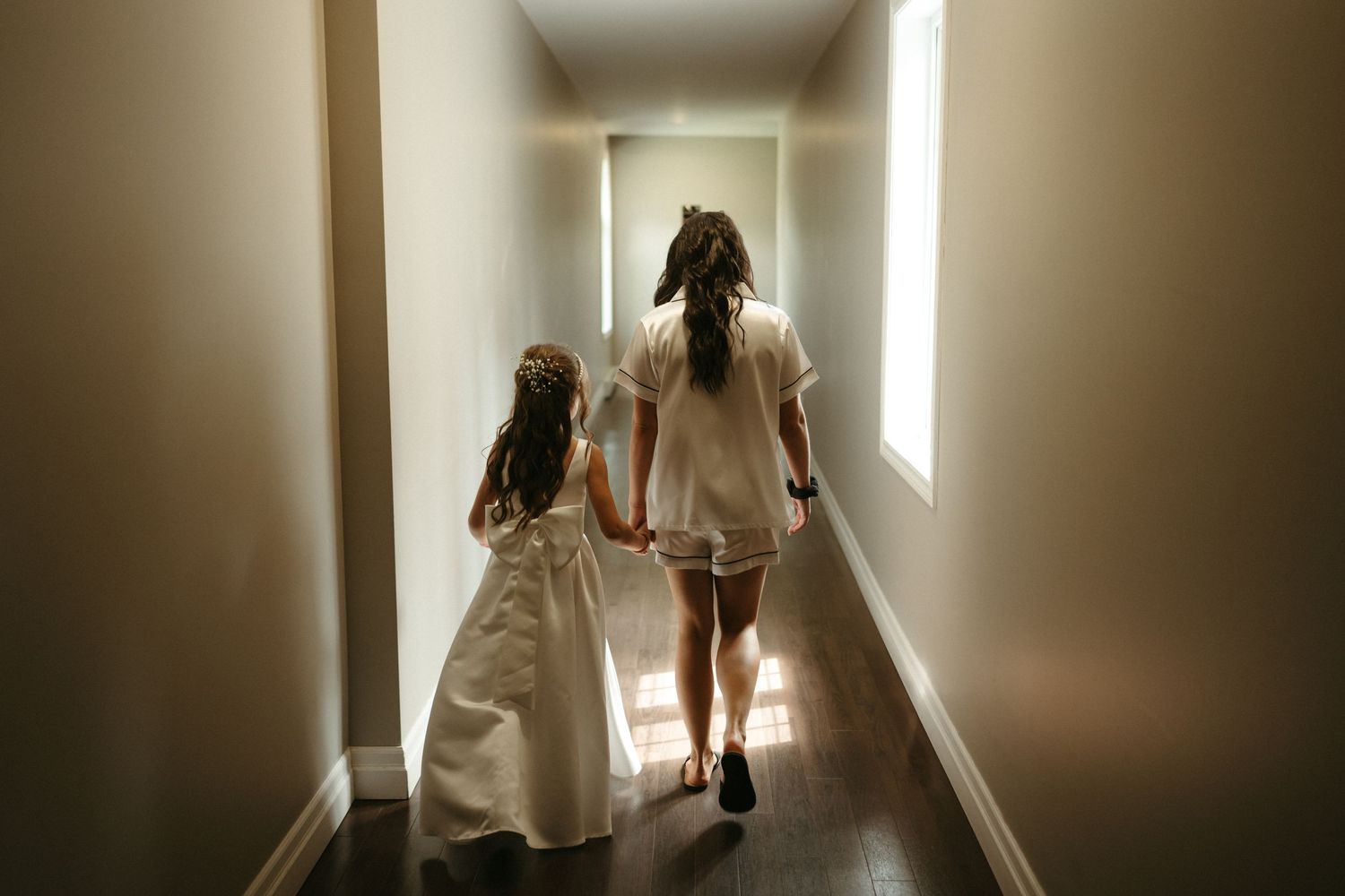 Bride and flower girl walking hand in hand down a softly lit hallway in white dresses.