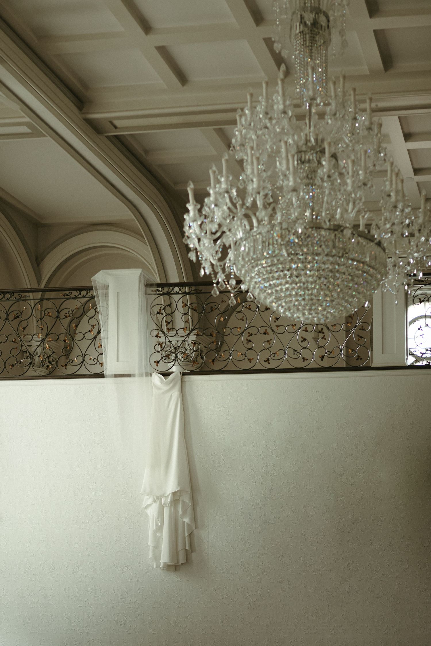 White wedding dress hanging near grand crystal chandelier in ornate venue with coffered ceiling and iron balcony.