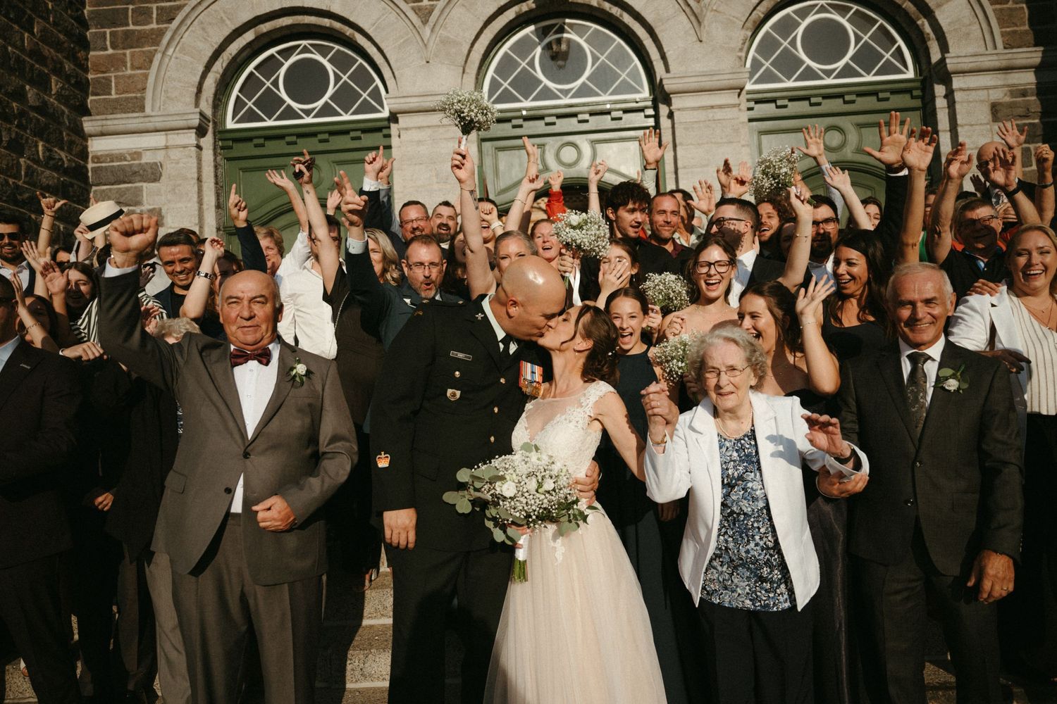 Newlyweds kiss on church steps surrounded by cheering wedding guests in sunny celebration.