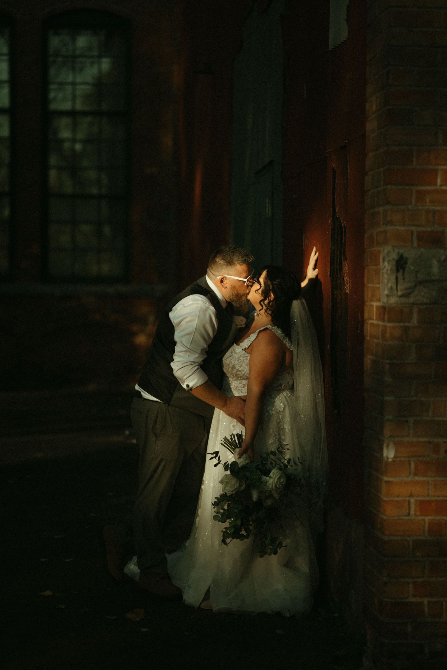 A couple in a romantic embrace against a dimly lit brick wall, the bride in a white gown and the groom in a suit.