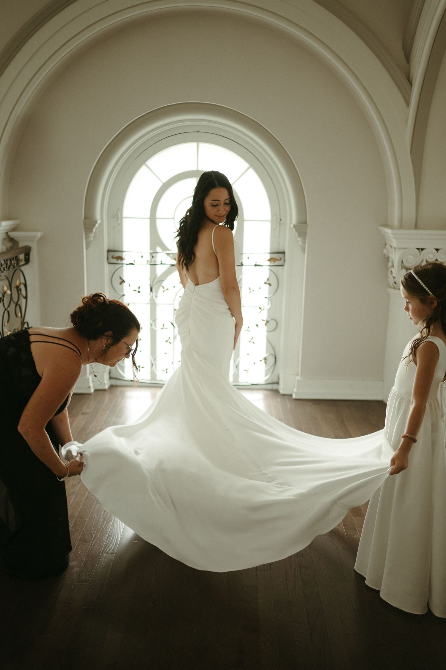 Bride in a white gown has her train arranged by helpers in an elegant arched foyer.