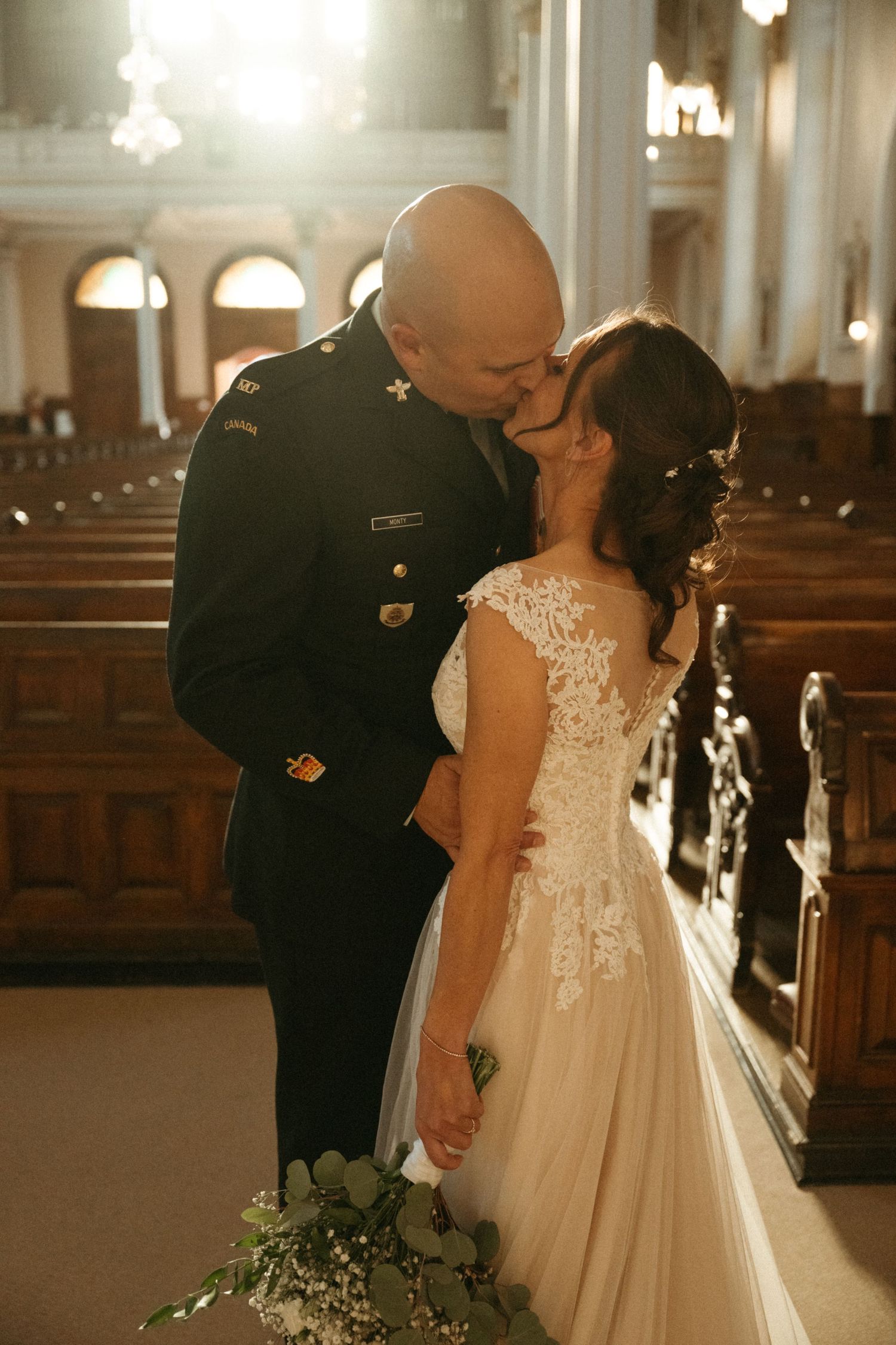 Military groom in black uniform shares first kiss with bride in lace gown inside sunlit church.