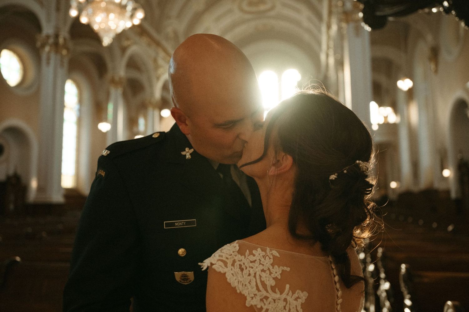 Military groom kissing bride in lace dress inside ornate church with warm golden light.