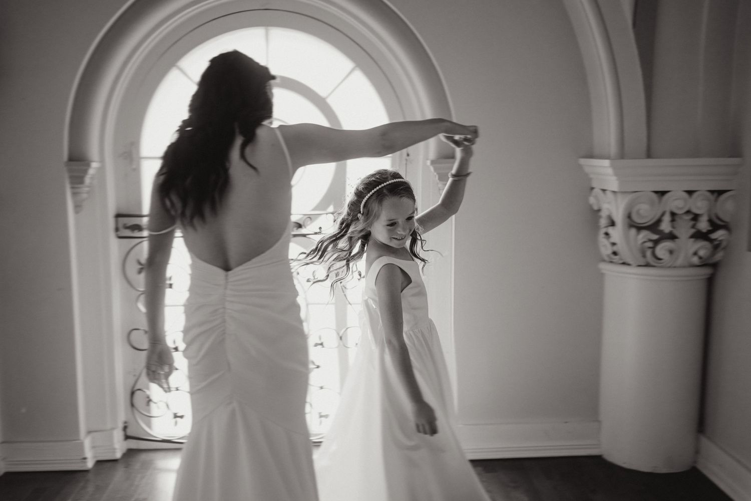 Bride in white gown twirling flower girl in elegant arched doorway, black and white wedding photo.