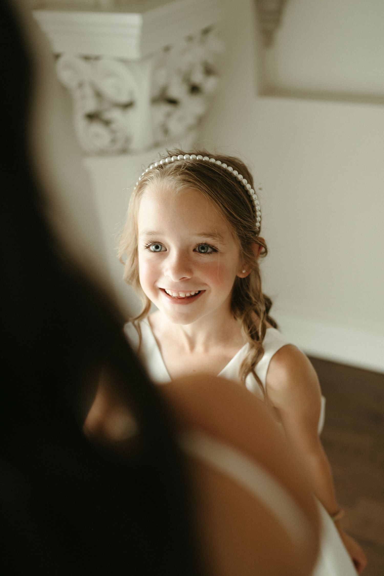 Young flower girl wearing pearl headband and white dress smiling warmly at wedding ceremony indoors.