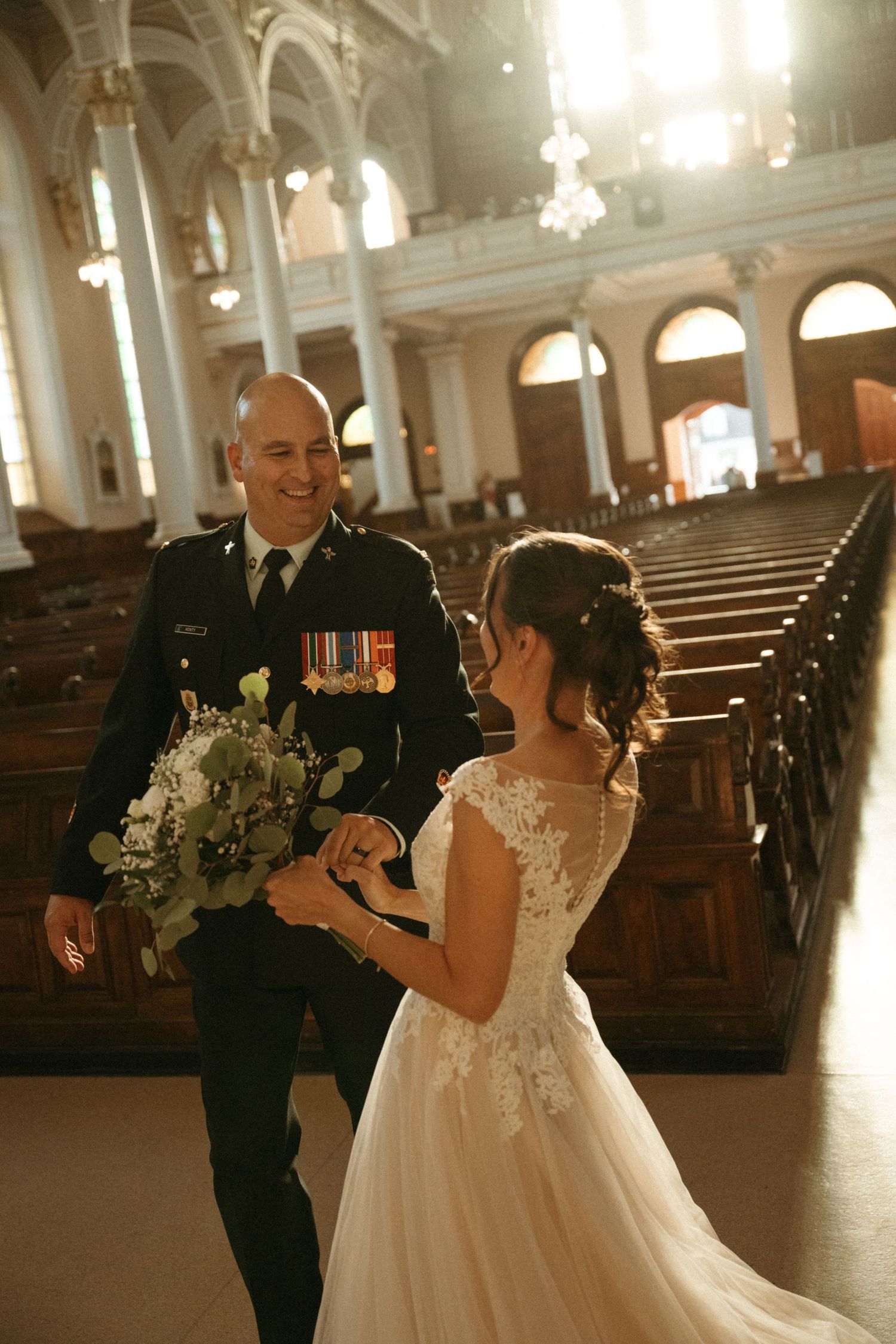 Military groom in uniform smiles at bride holding bouquet inside stunning historic church.