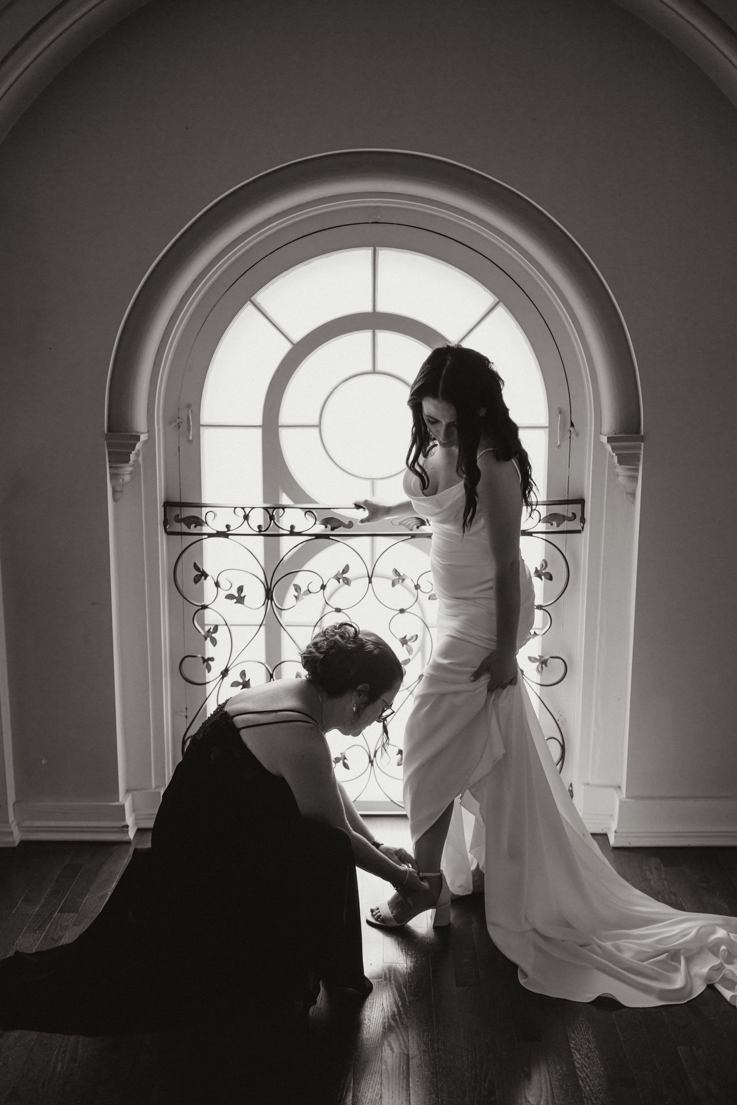 Bride in white gown getting shoes fastened by bridesmaid in black dress before arched window, black and white photo.