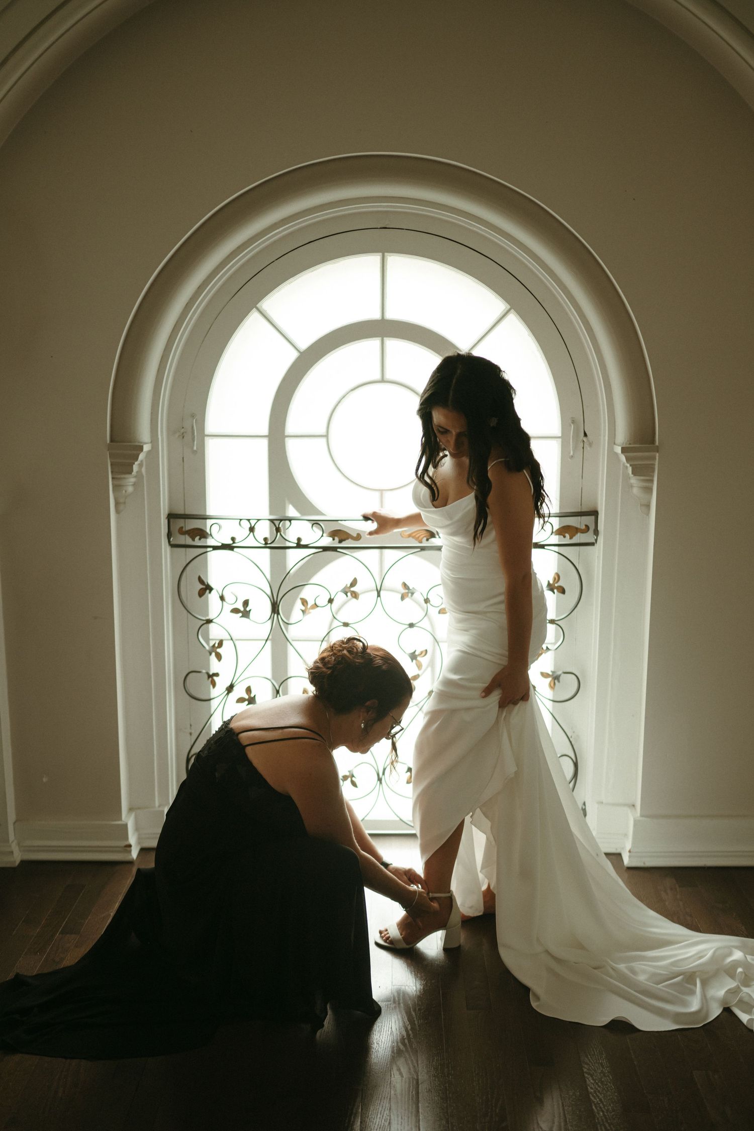 Bridesmaid kneeling to help bride with shoes in front of stunning arched window with iron railing.