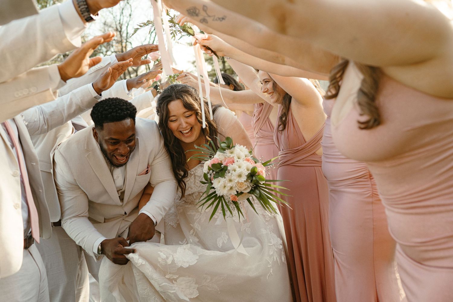 Bride and groom ducking under arch of hands made by wedding party during outdoor celebration.
