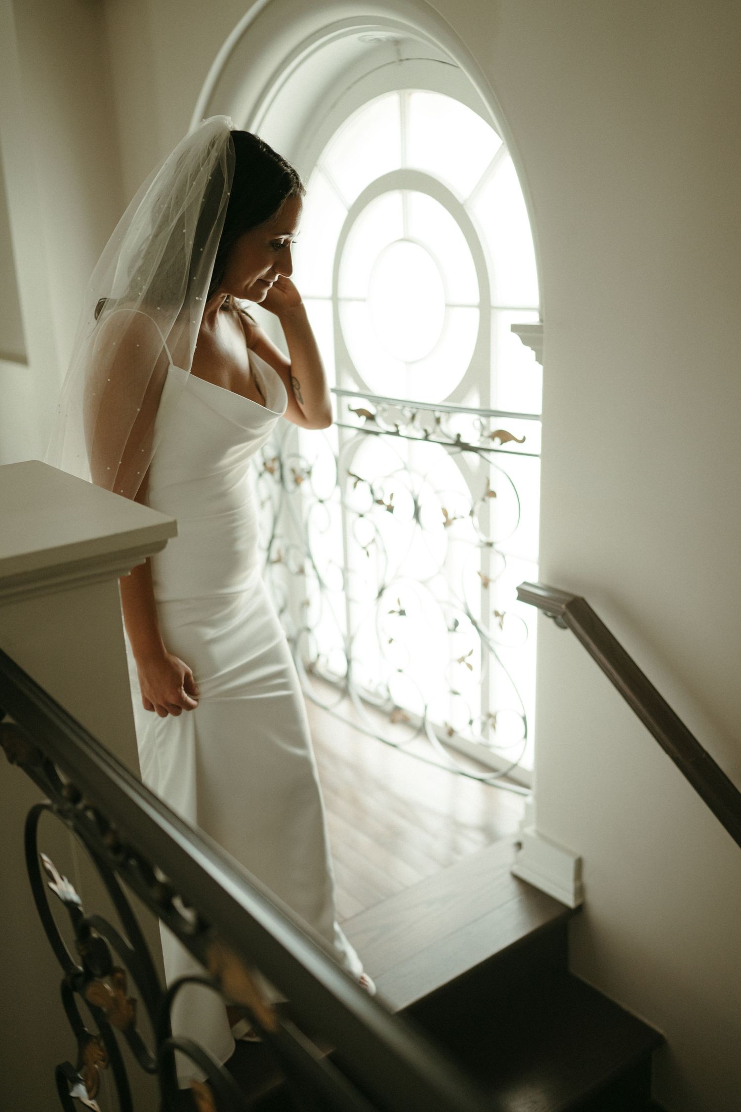 Elegant bride in white gown and veil standing on staircase beside ornate arched window.