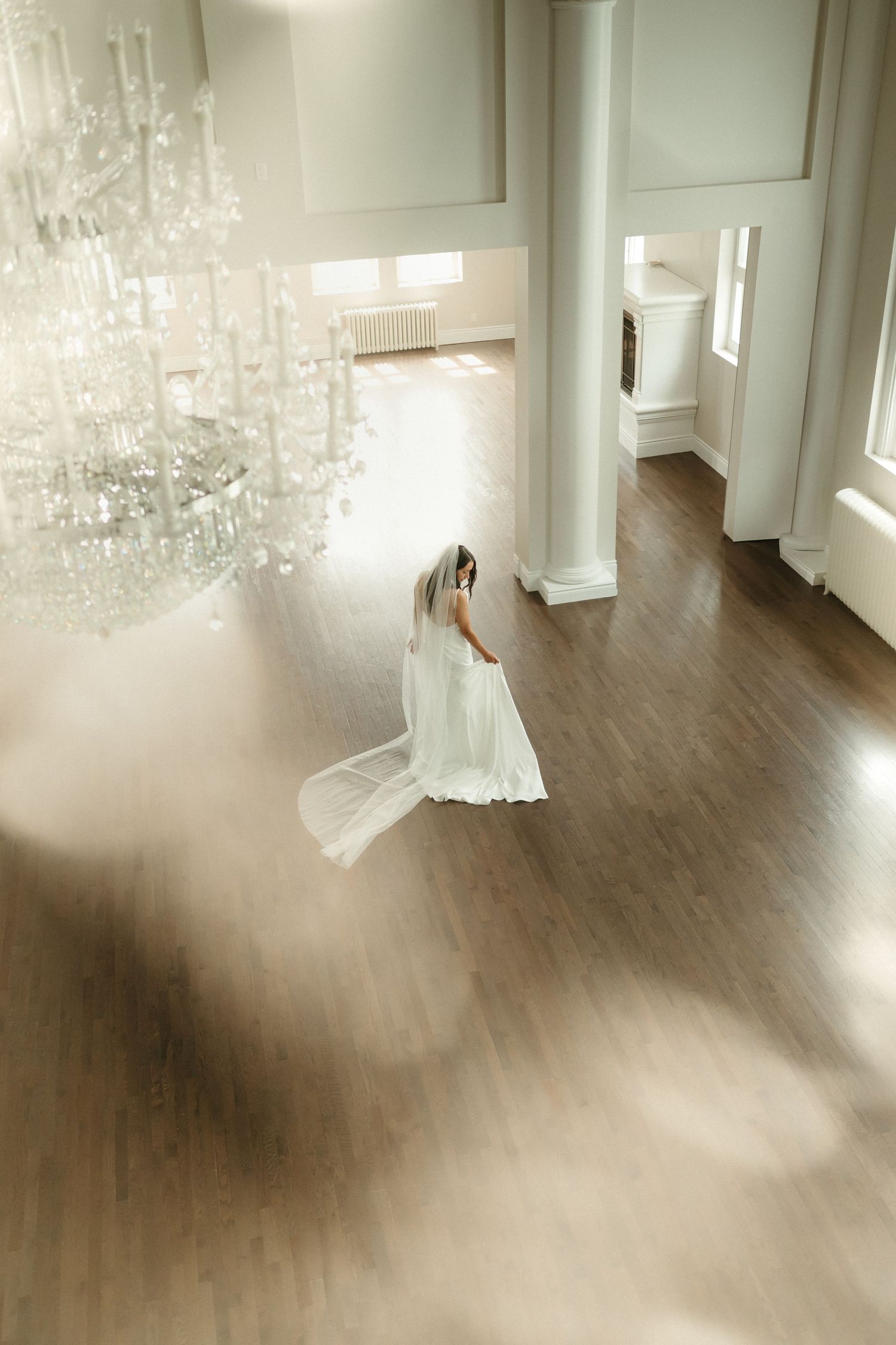 Bride in white gown and veil walks through elegant bright ballroom beneath crystal chandelier.